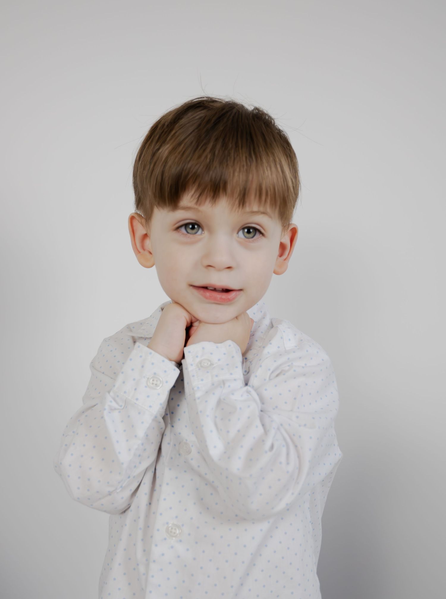 A young child in a white lace shirt poses sweetly with hands under chin.