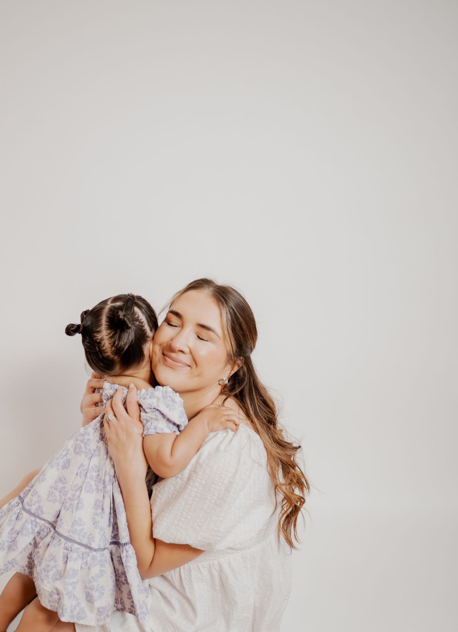 Tender embrace between parent and child in matching white dresses against neutral background.