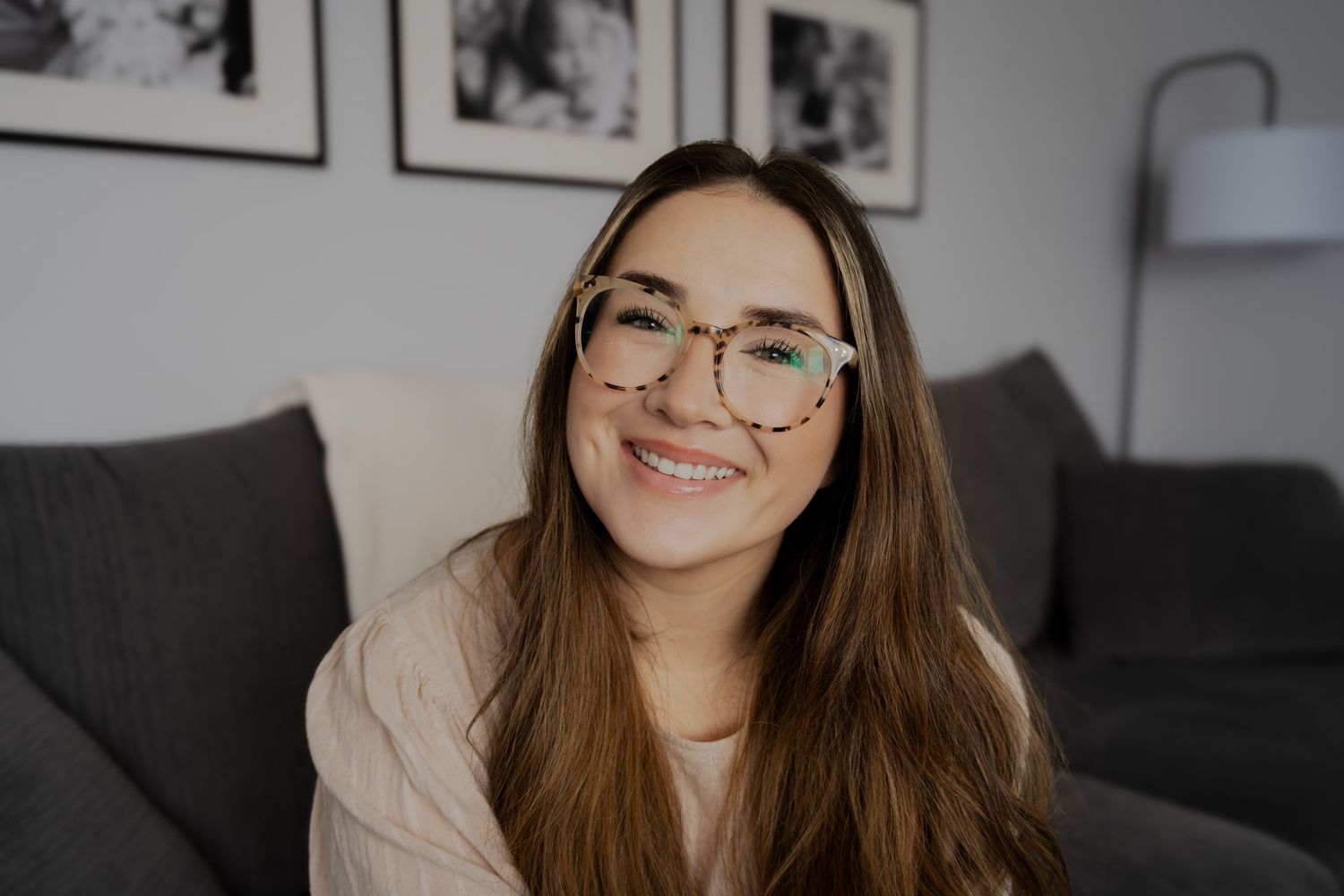Candid portrait of woman with glasses and long hair laughing on grey couch.