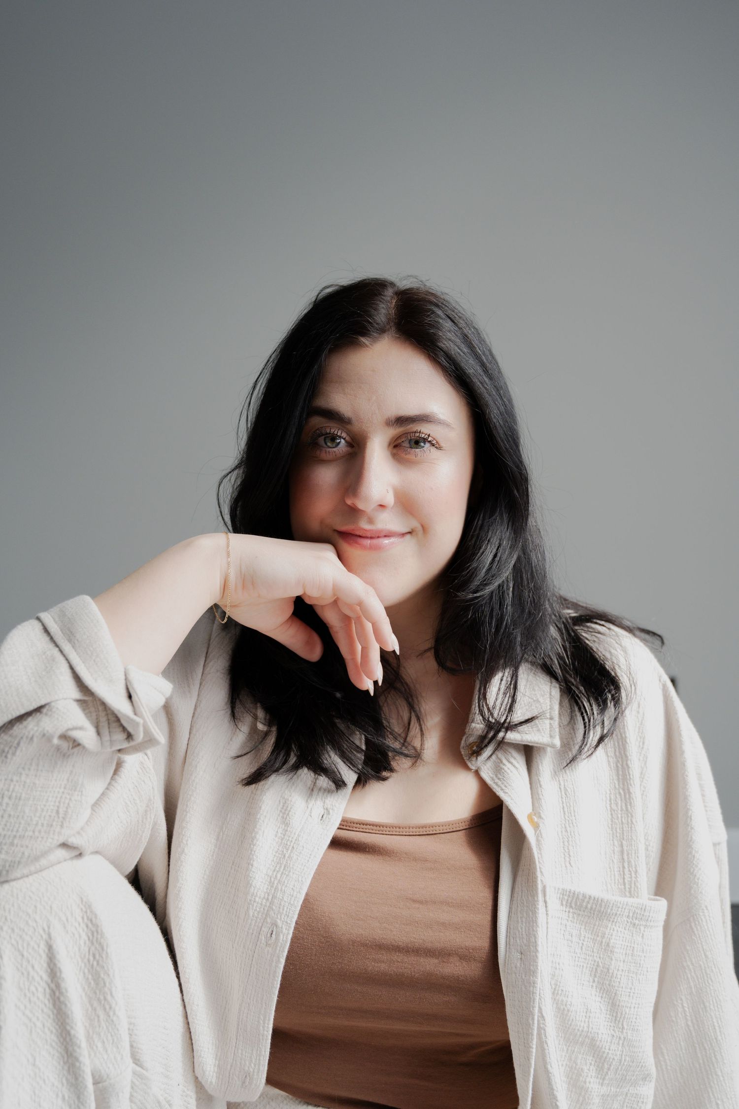 Portrait of a person in a beige jacket and brown top against a gray background with a gentle smile.