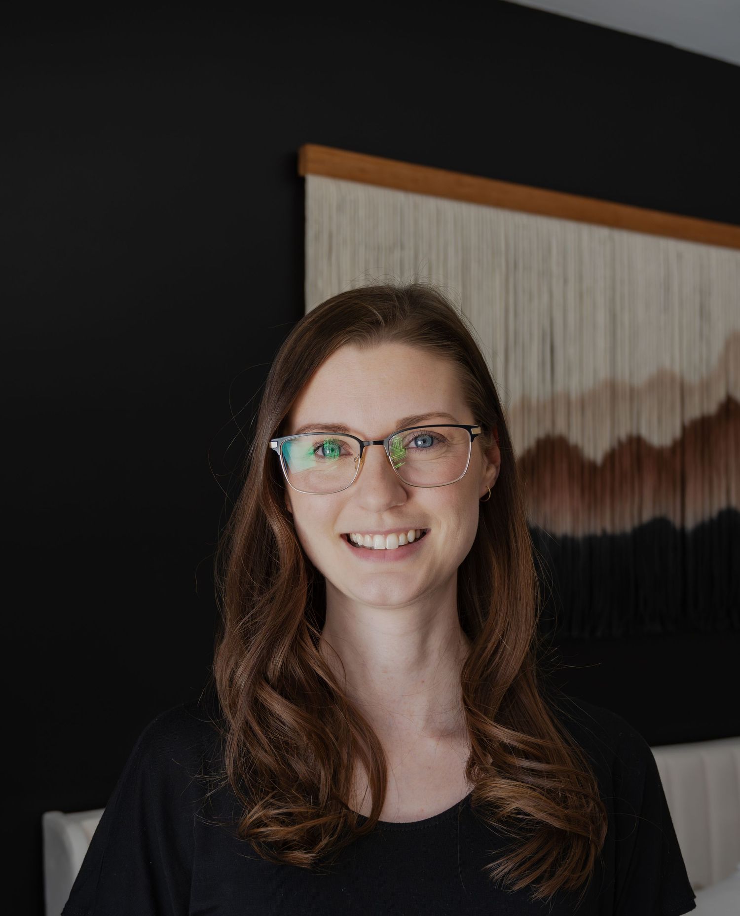 Portrait against dark wall with artistic wall hanging in background.