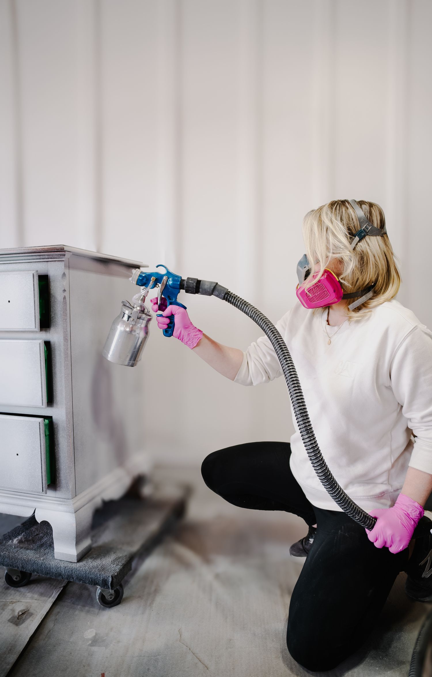 Person using paint sprayer on furniture while wearing protective mask and gloves.