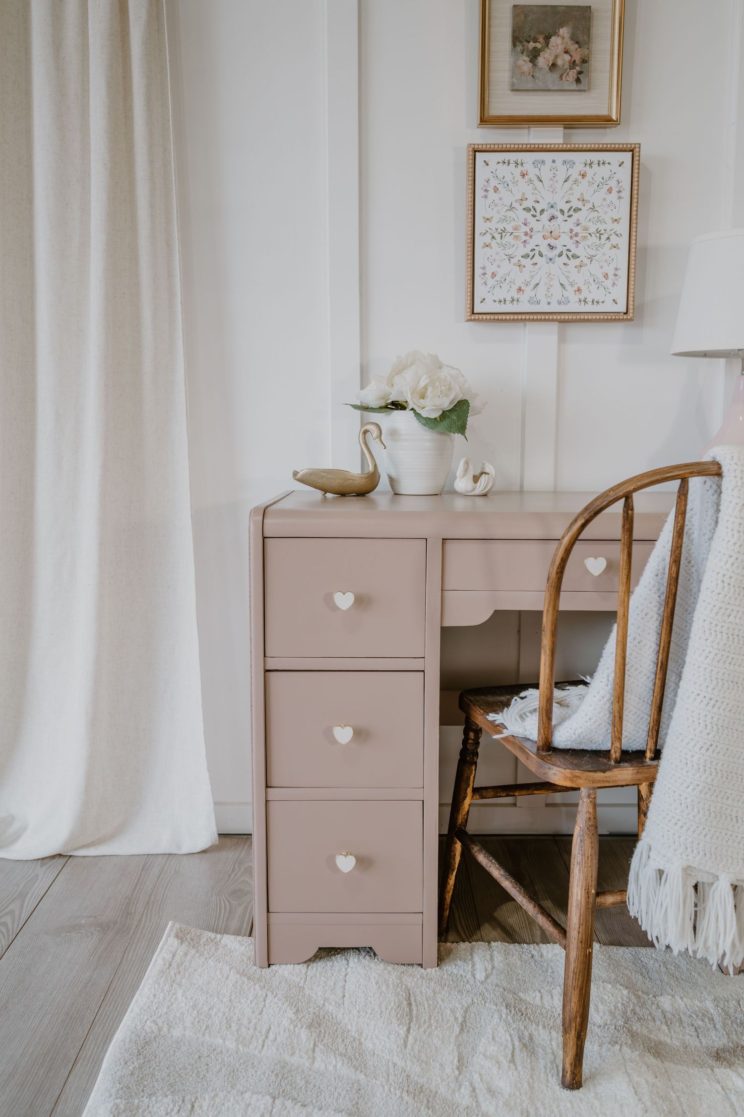 Vintage-style desk in beige with white heart knobs beside wooden chair and framed wall art.