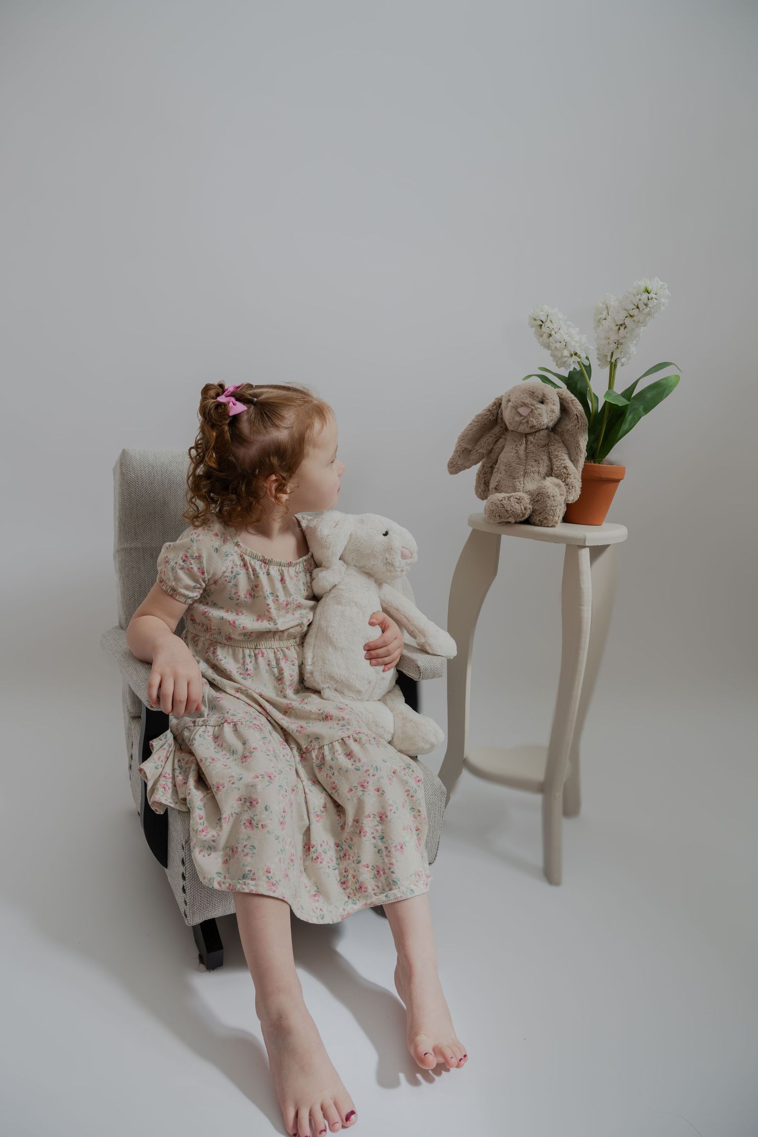 Child in vintage floral dress sits with stuffed bunny toy on chair next to side table with flowers.