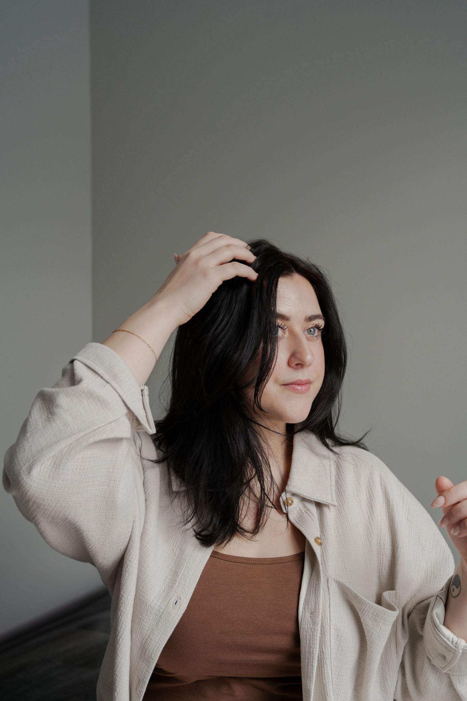 Portrait of someone in beige jacket running hand through dark hair against grey background.