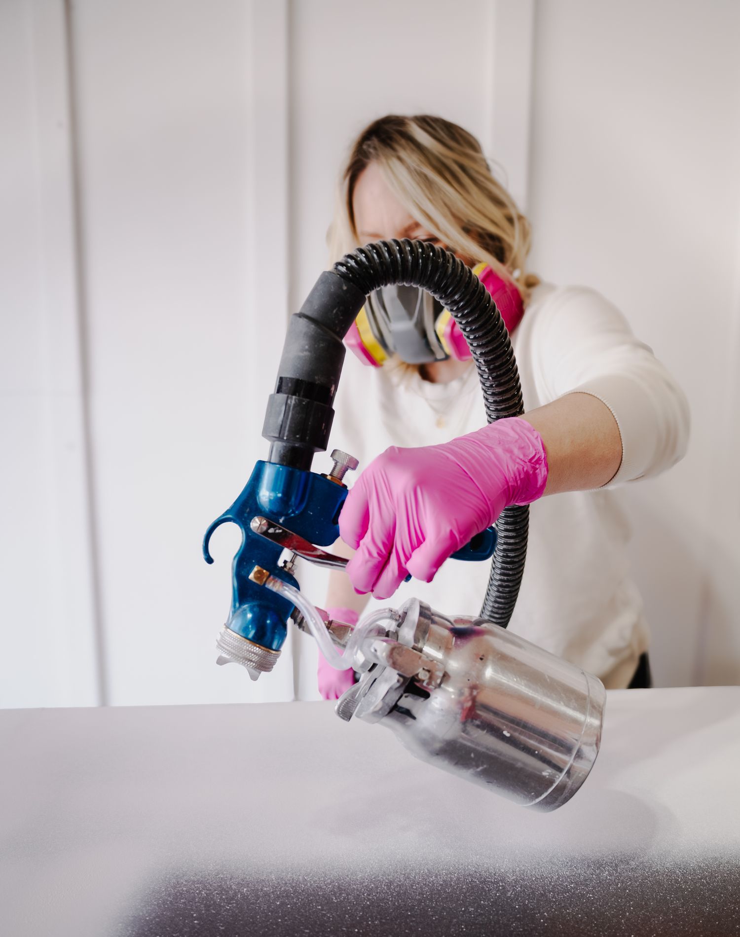 Person wearing protective gear uses paint sprayer with pink gloves and white long sleeves.