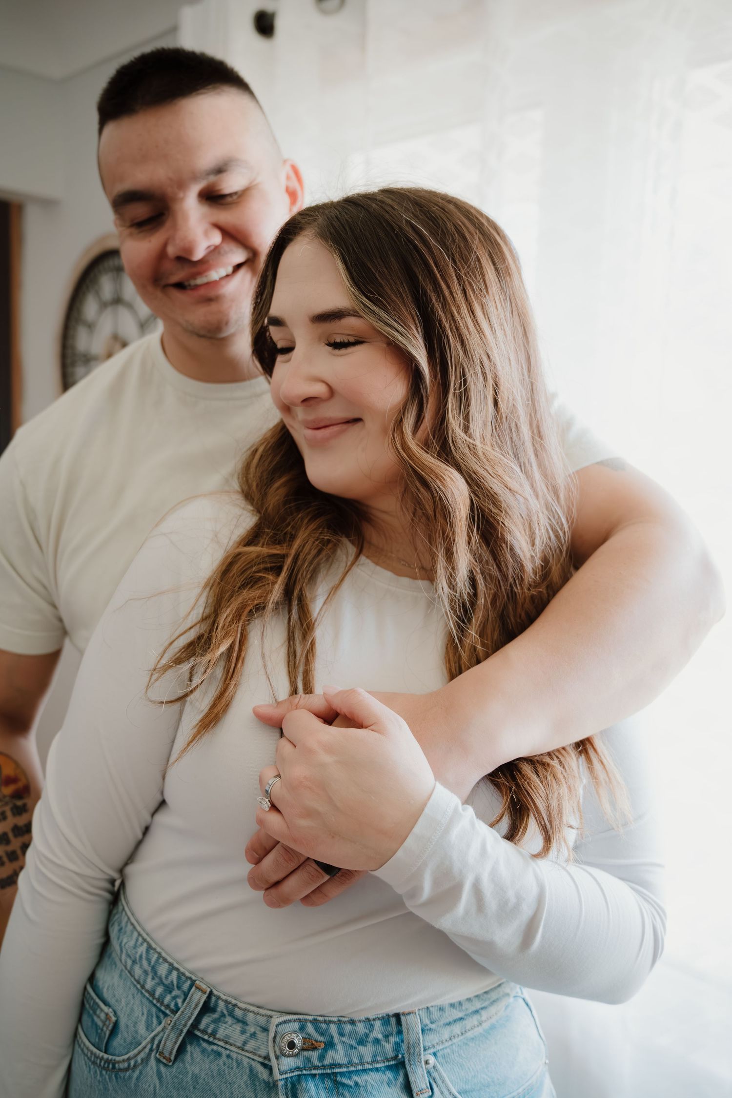 Couple embracing in casual attire against white wall with clock.