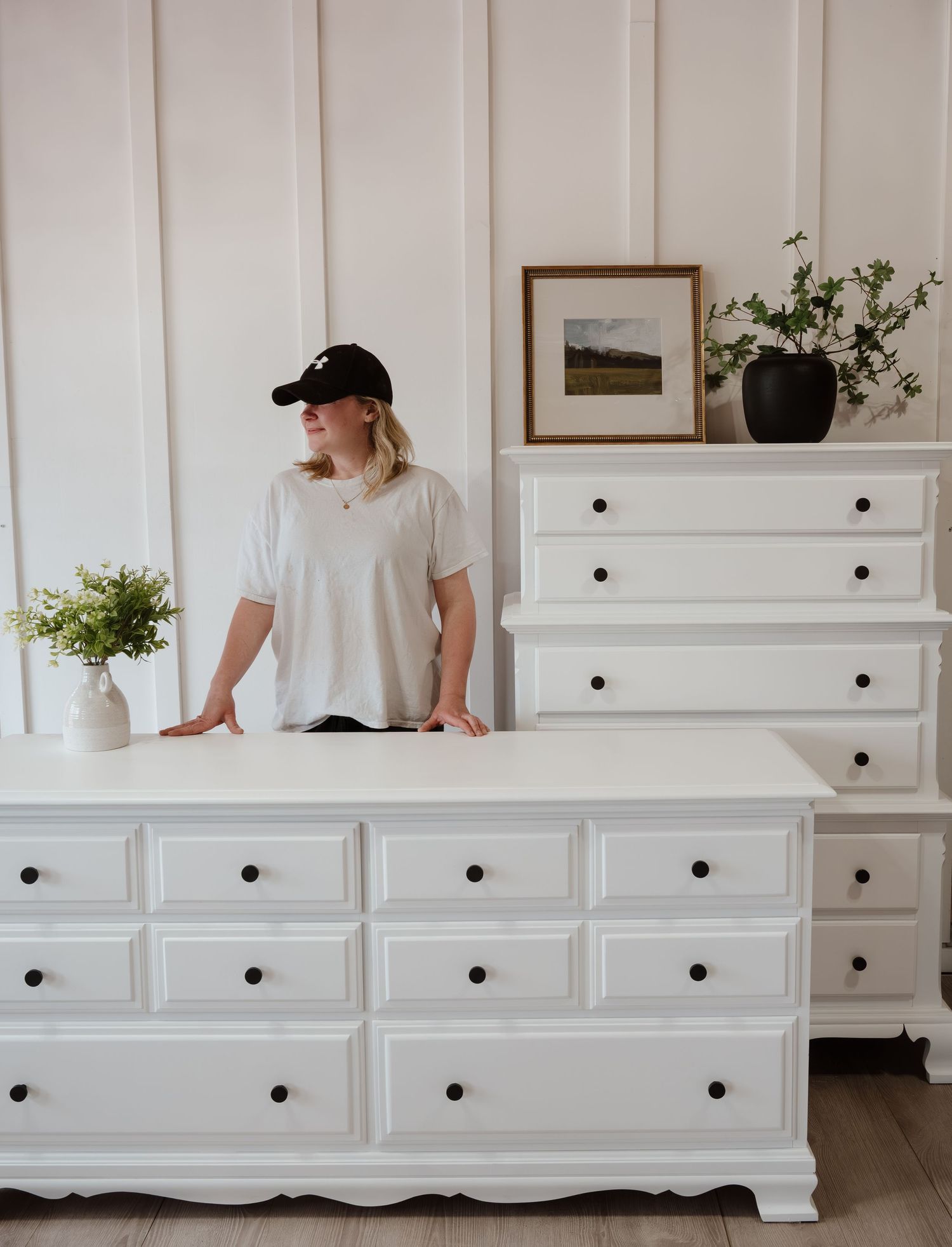 White painted dresser displayed against paneled wall with decorative plants and artwork.