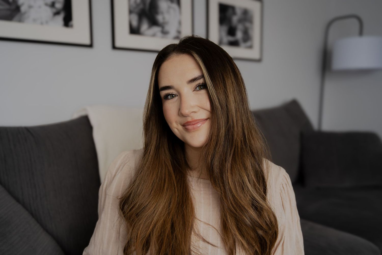 Warm indoor portrait of woman with long hair smiling on grey couch.