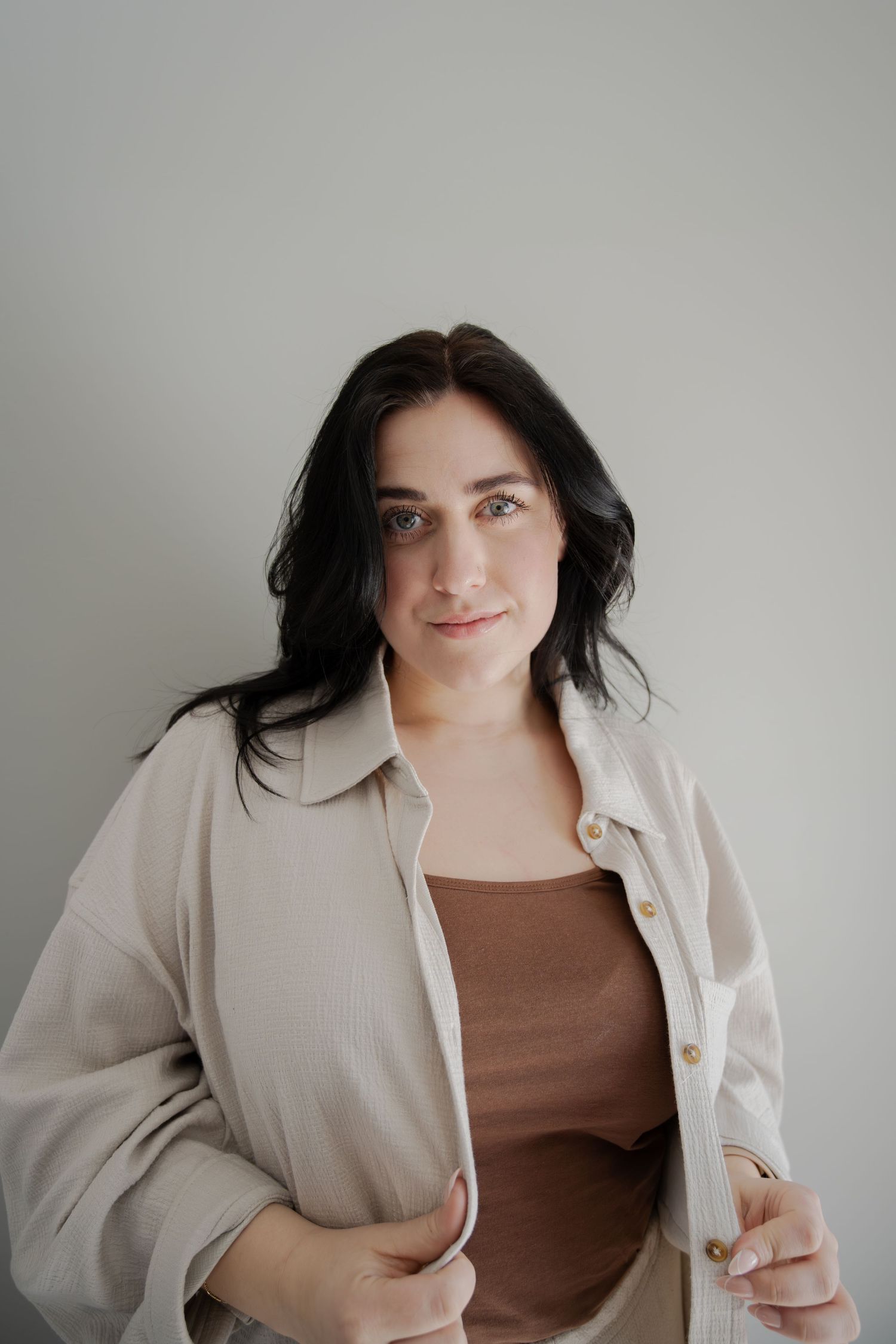 Fashion portrait of woman in cream jacket and brown top against minimal grey background.