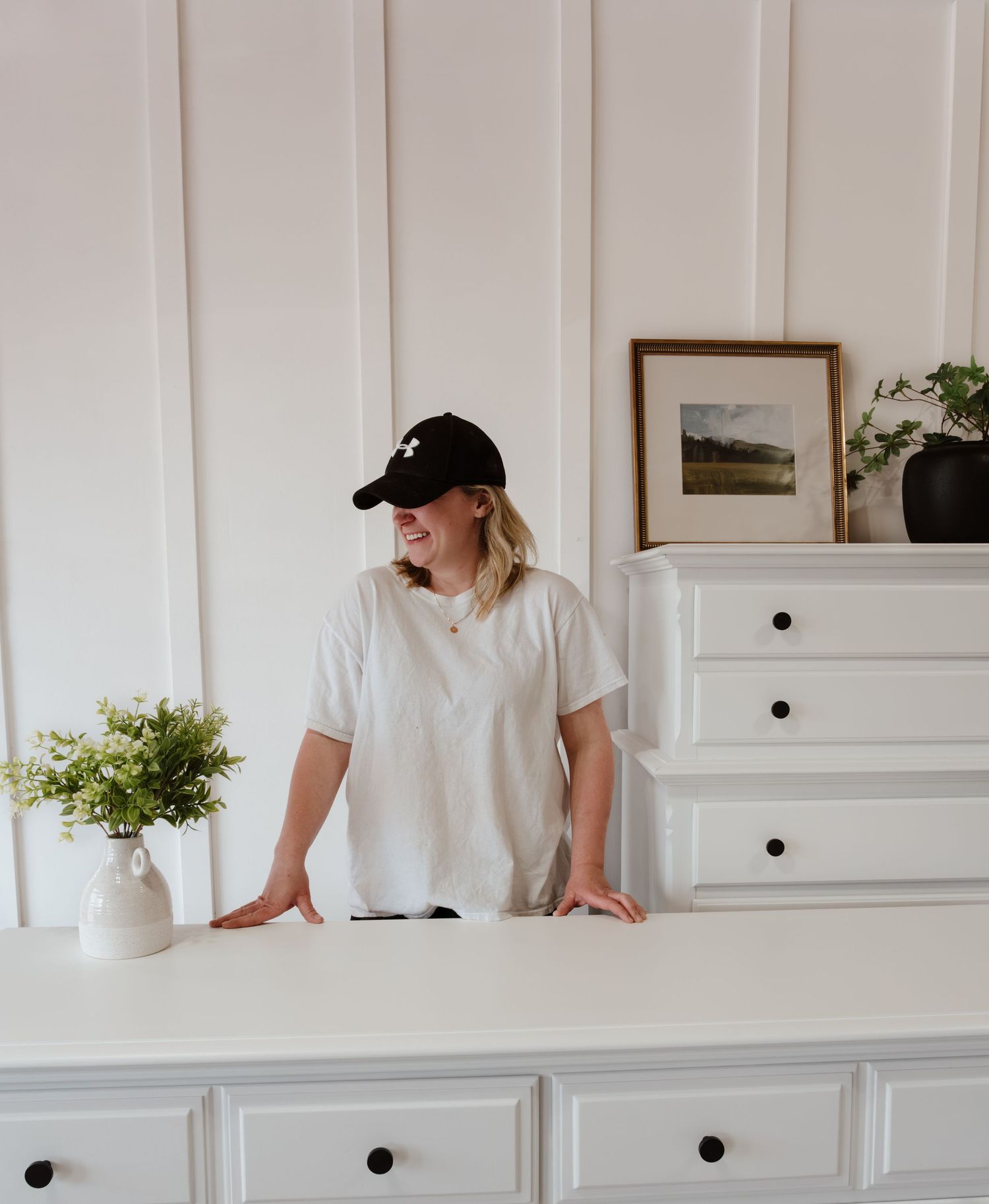 Person in casual attire standing at a white dresser with decorative plants and artwork.