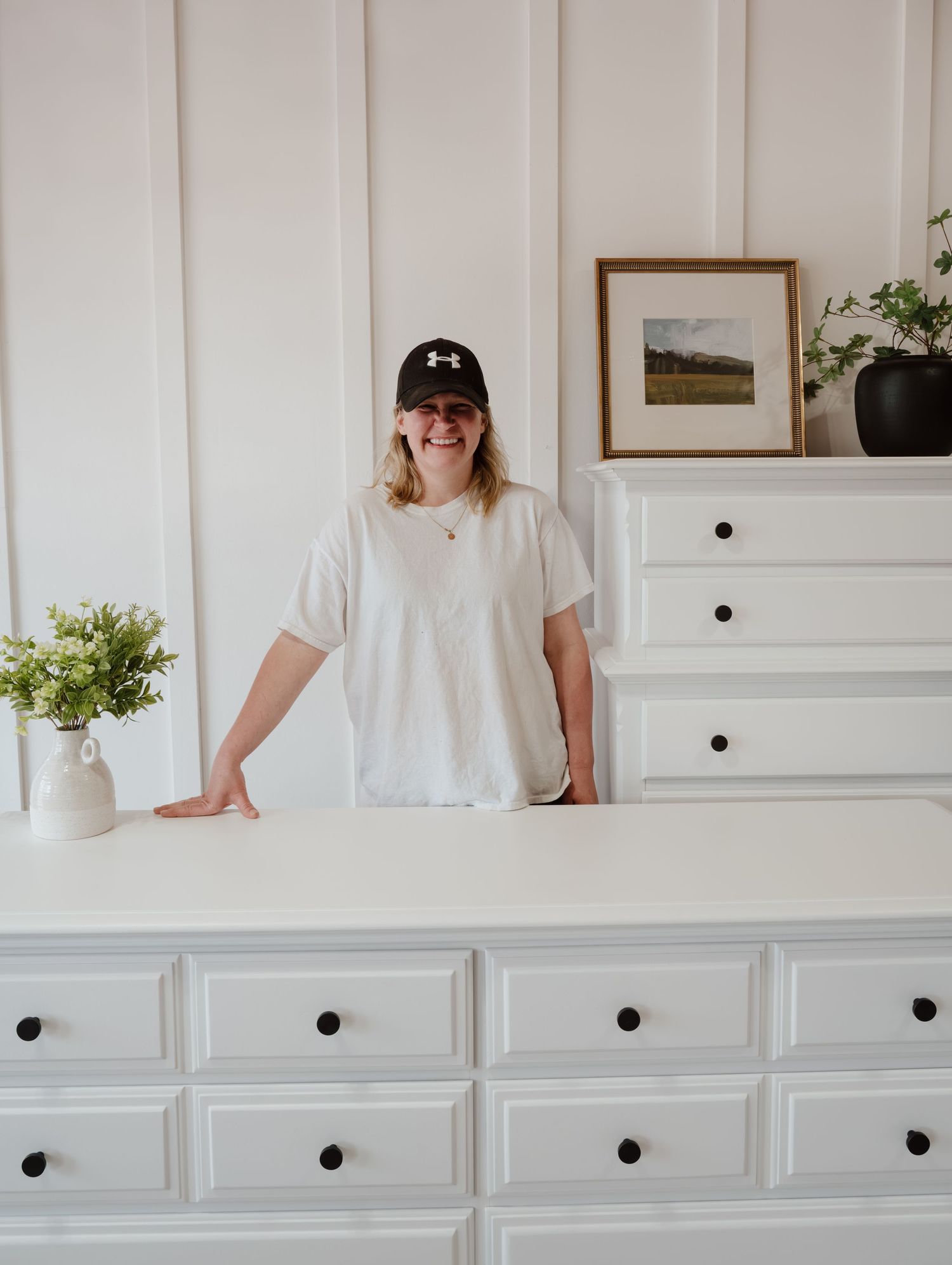 Person in white shirt and baseball cap stands beside white dresser in bright minimalist room.