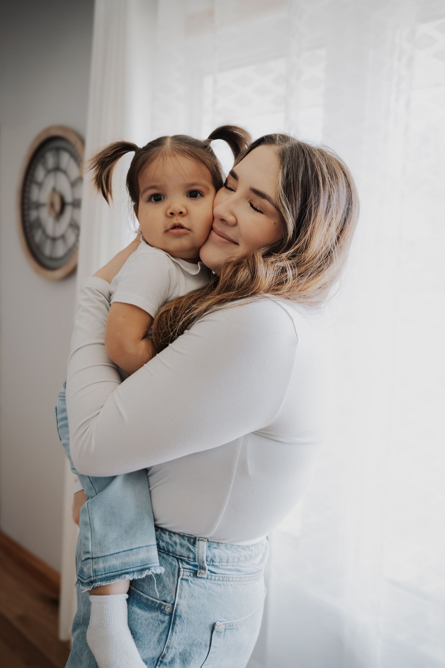 Intimate moment between parent and child in casual wear against white wall with clock.
