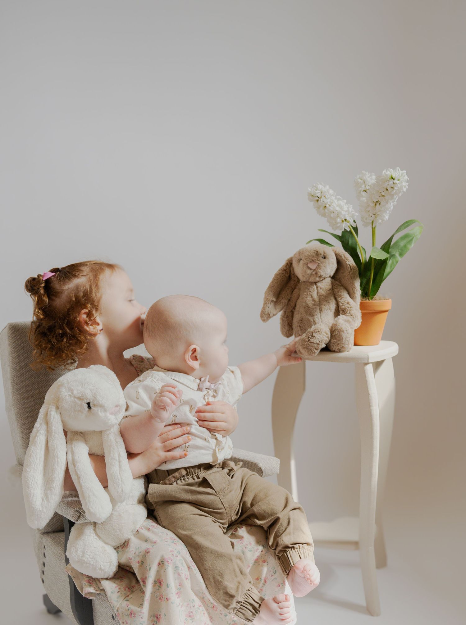 Children looking at white flowers on a side table with plush bunnies in a minimalist setting.