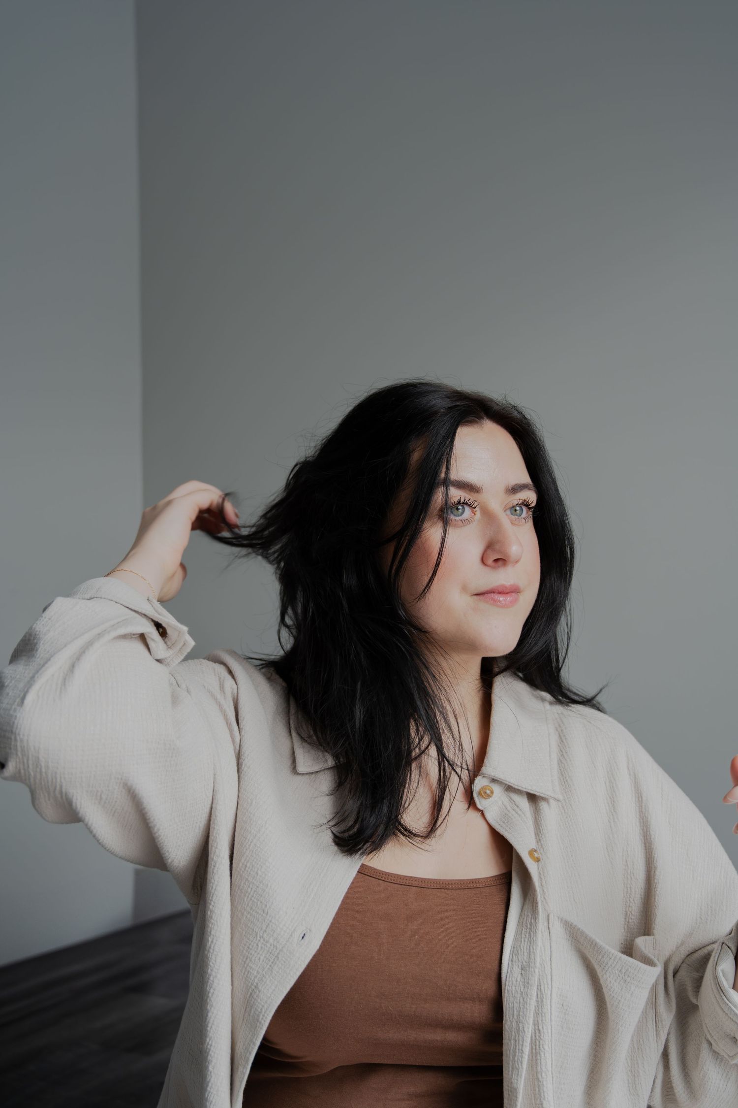 Woman in beige jacket and brown top playing with dark hair against grey wall.