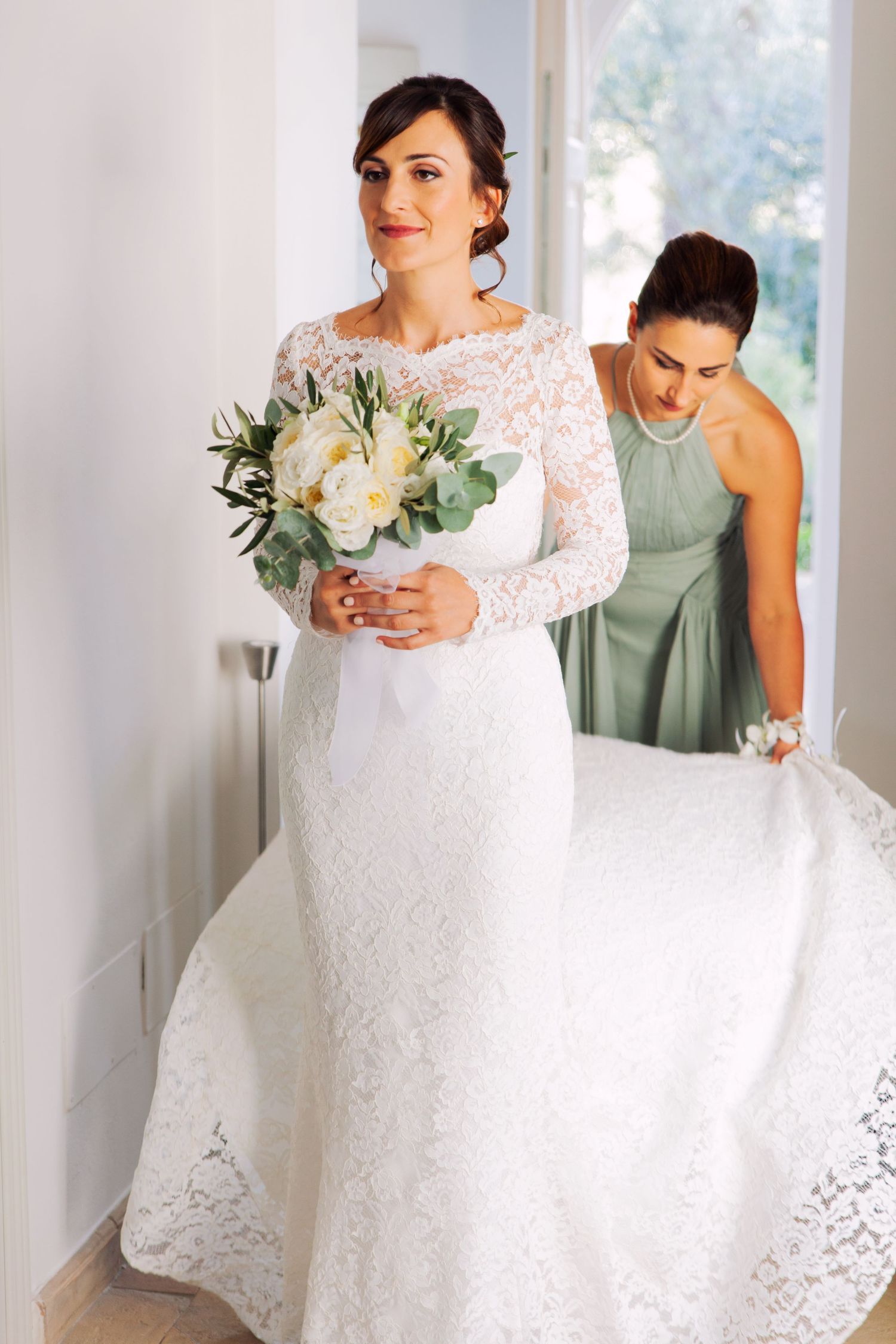 Smiling bride in lace wedding dress holding white bouquet while bridesmaid adjusts train.