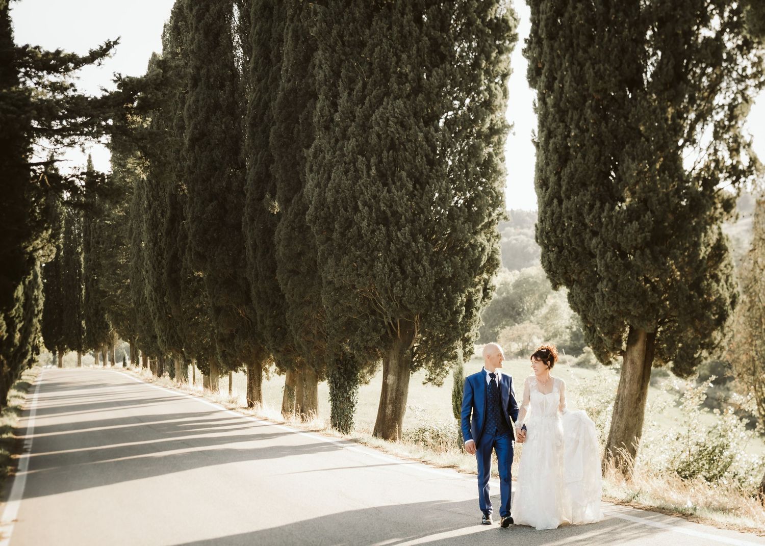 Bride and groom walk hand in hand along a cypress tree-lined road in the Tuscan countryside.
