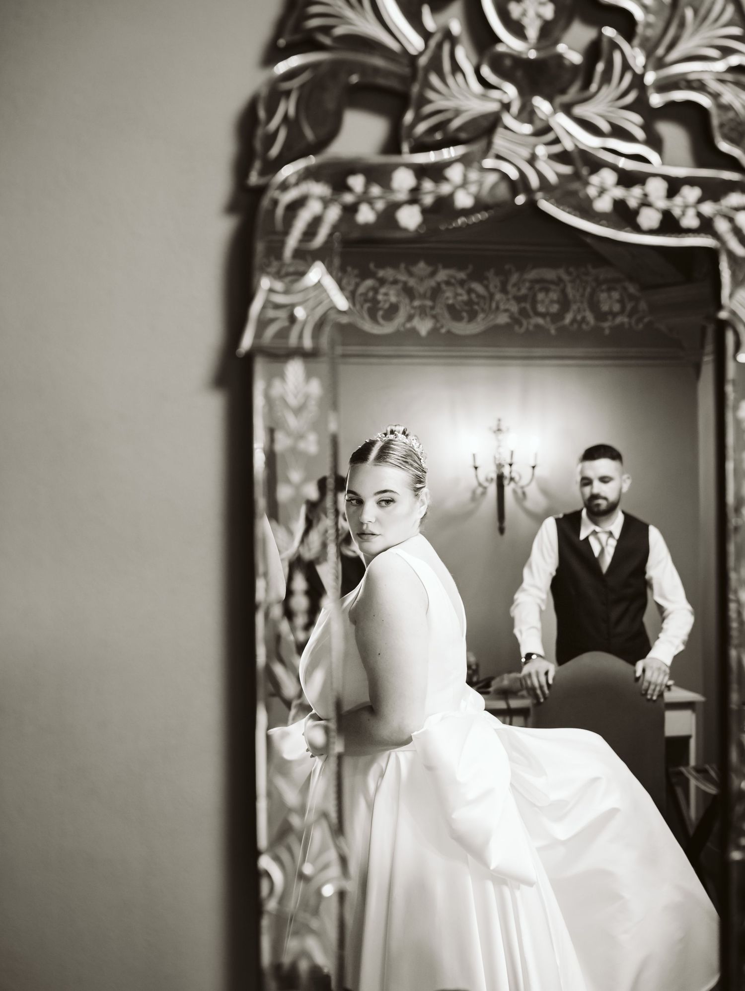 Black and white mirror reflection of bride in white gown with groom standing behind.