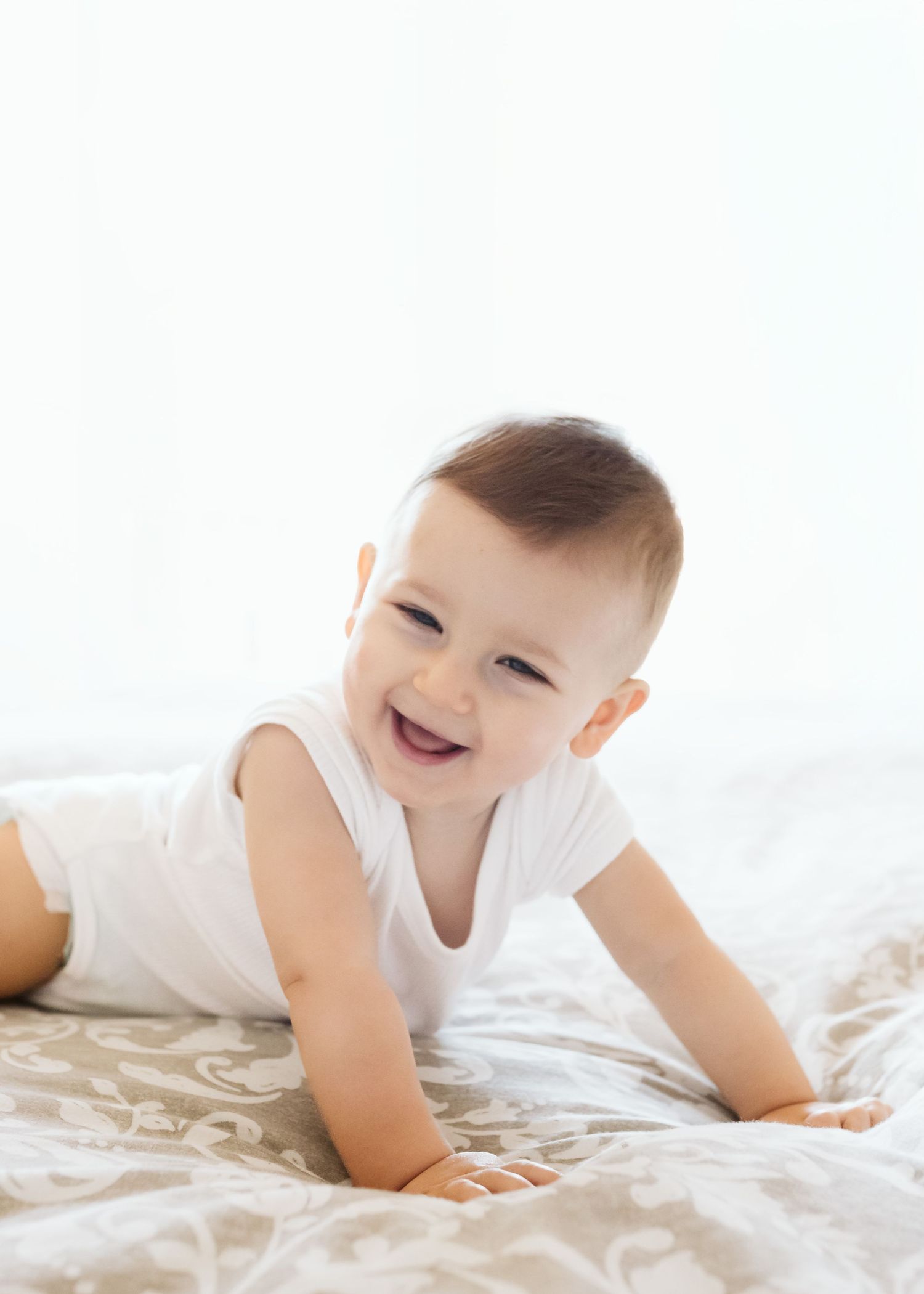 Happy smiling baby in white onesie doing tummy time on patterned bedding.