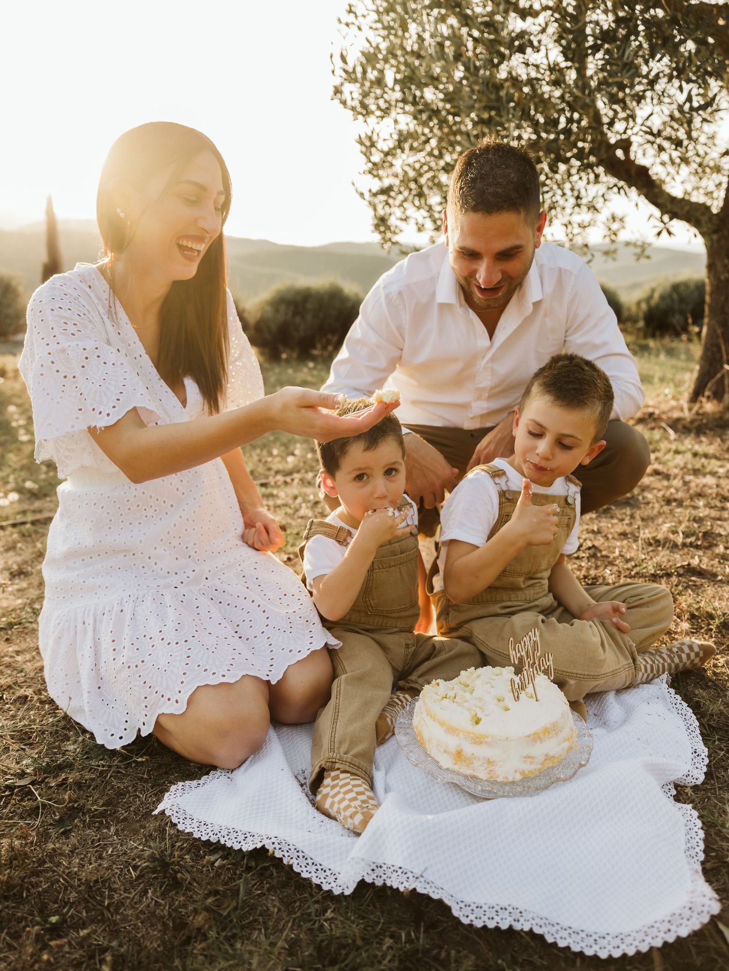 Happy family celebrating toddlers birthday with cake outdoors in golden sunset light.