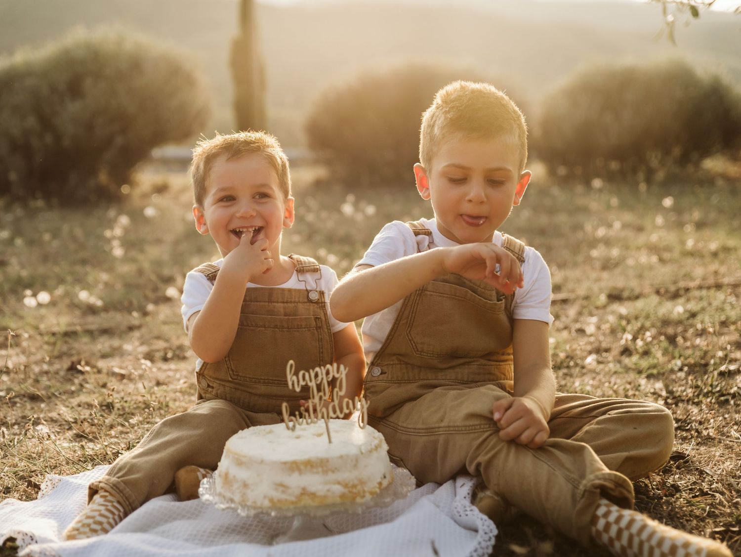 Two young twin boys in brown overalls celebrating birthday, eating cake outdoors in golden sunlight.