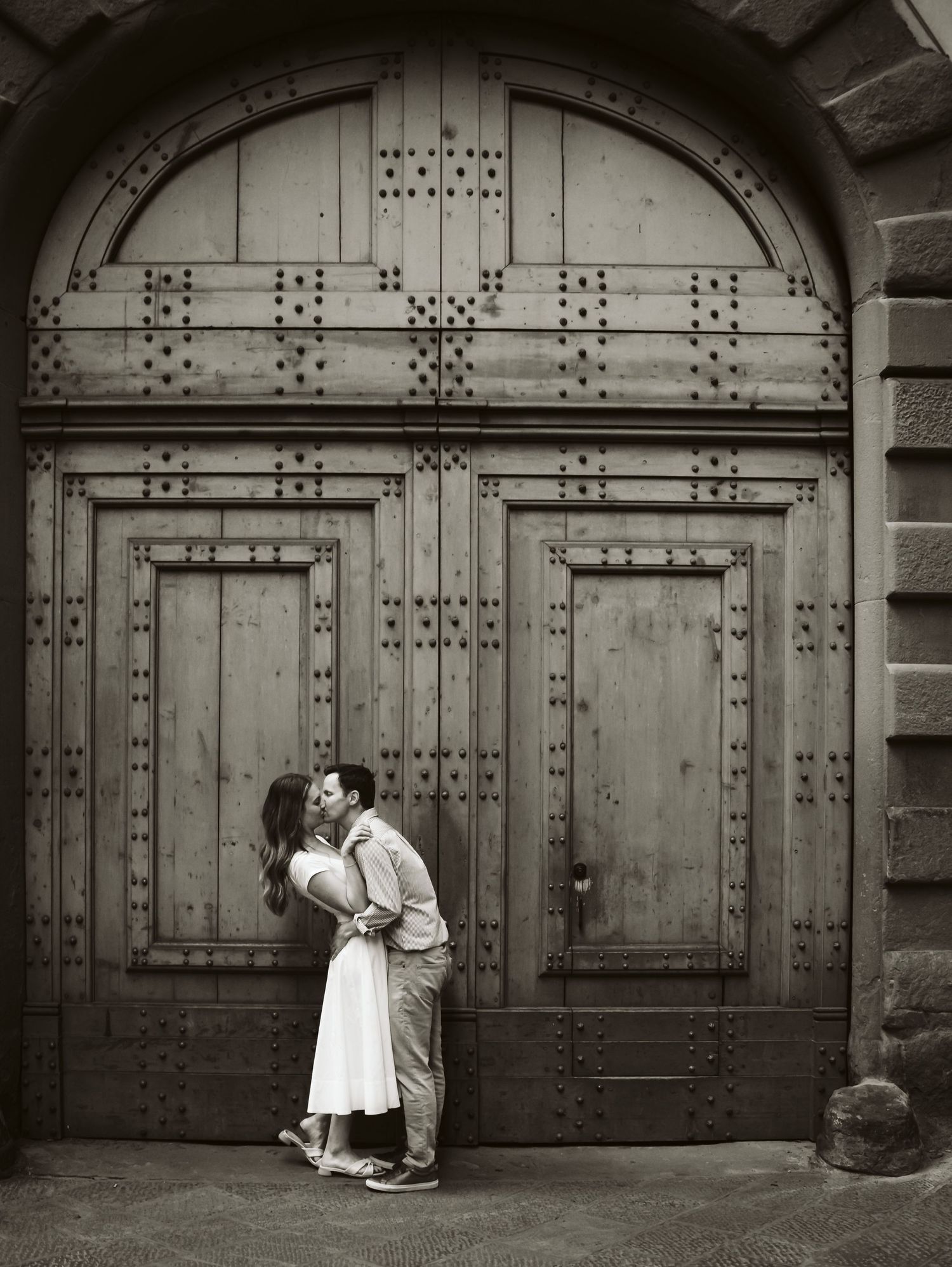Couple sharing a kiss in front of a grand wooden door in black and white.