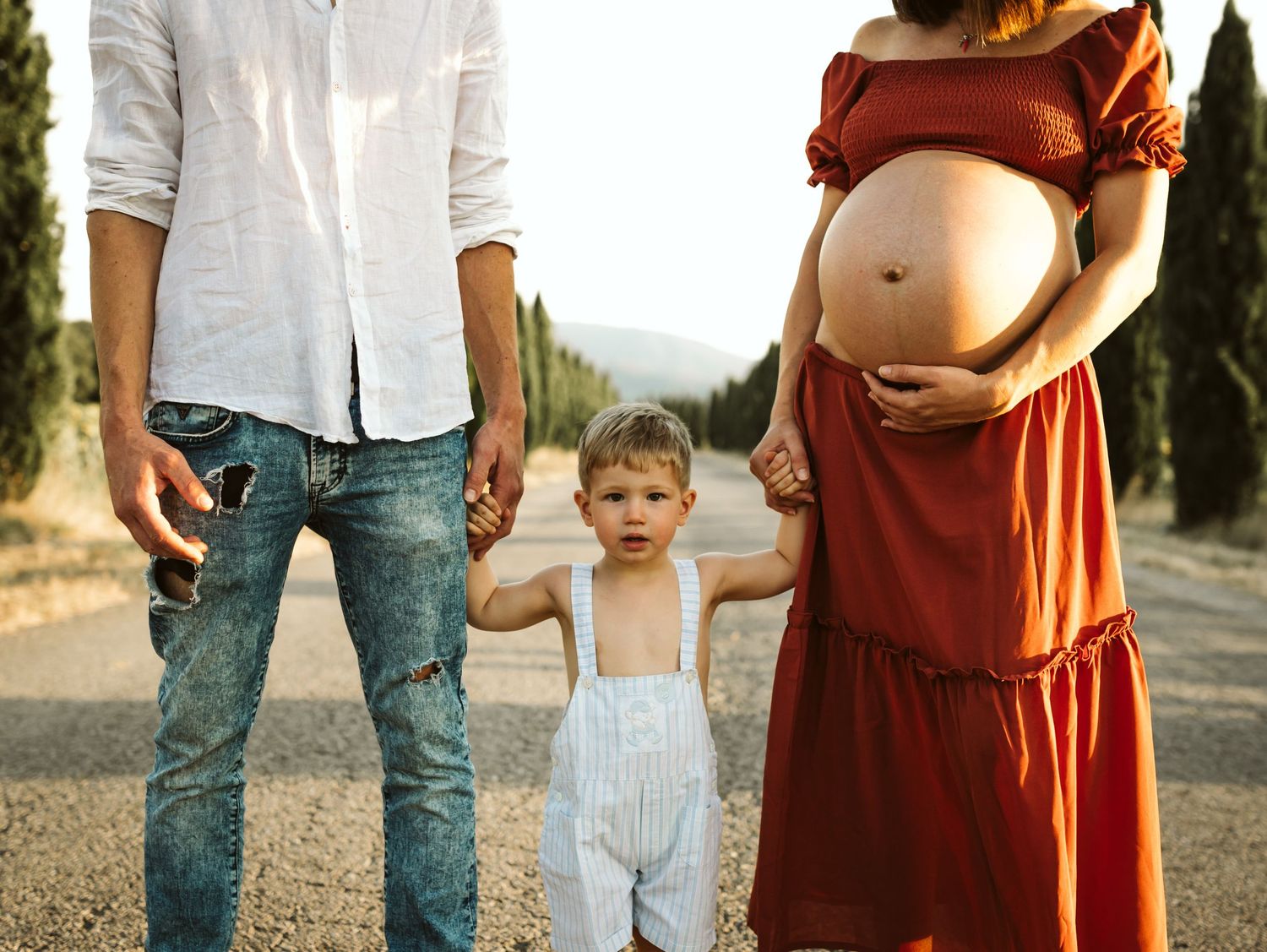 Pregnant woman in red dress and man holding hands with toddler boy on a sunny outdoor road.
