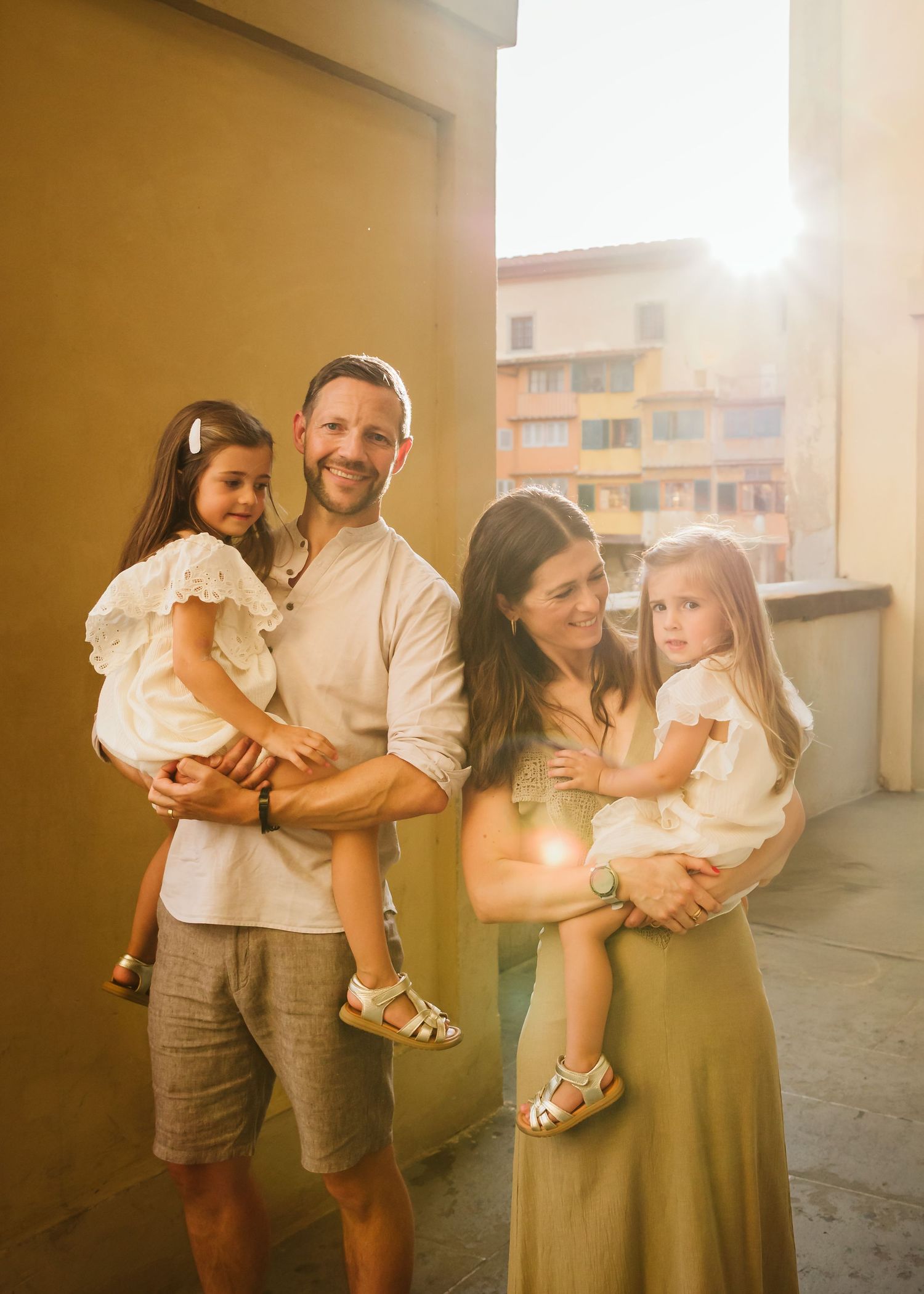 Smiling family of four bathed in golden sunlight, parents holding two young daughters outdoors.