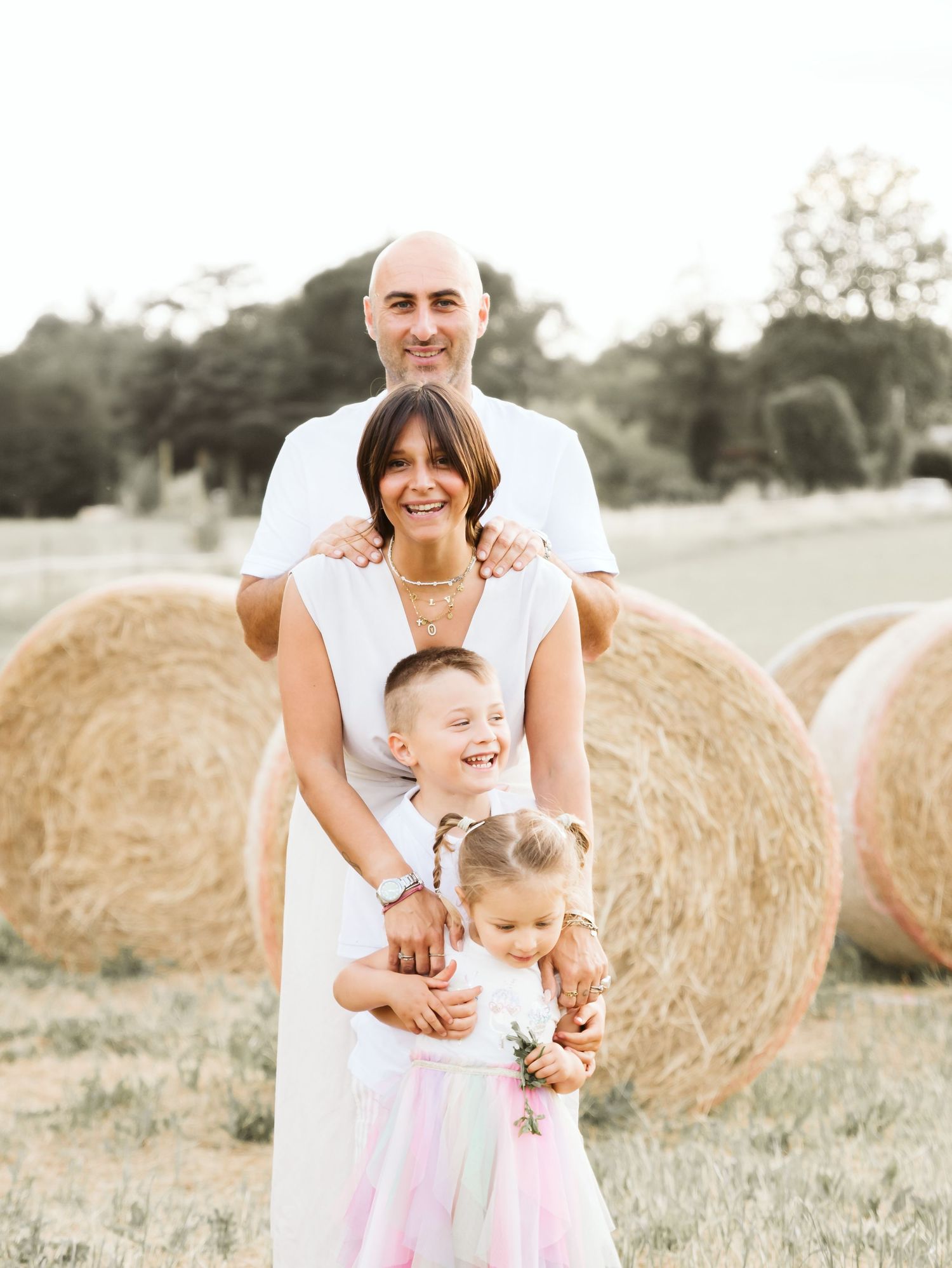 Family of four posing in a golden field with hay bales, parents embracing two young children in white outfits.