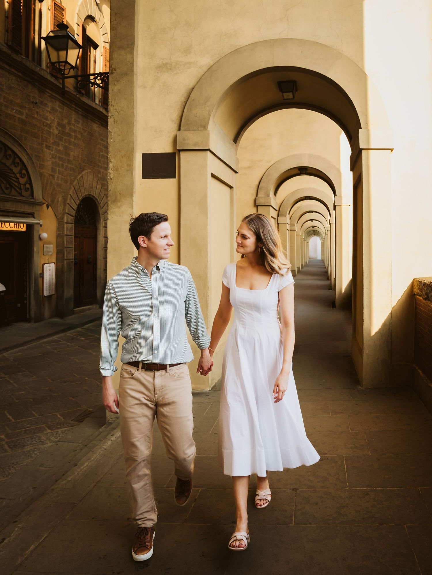 Couple strolling hand in hand through a golden archway corridor in Florence, Italy.