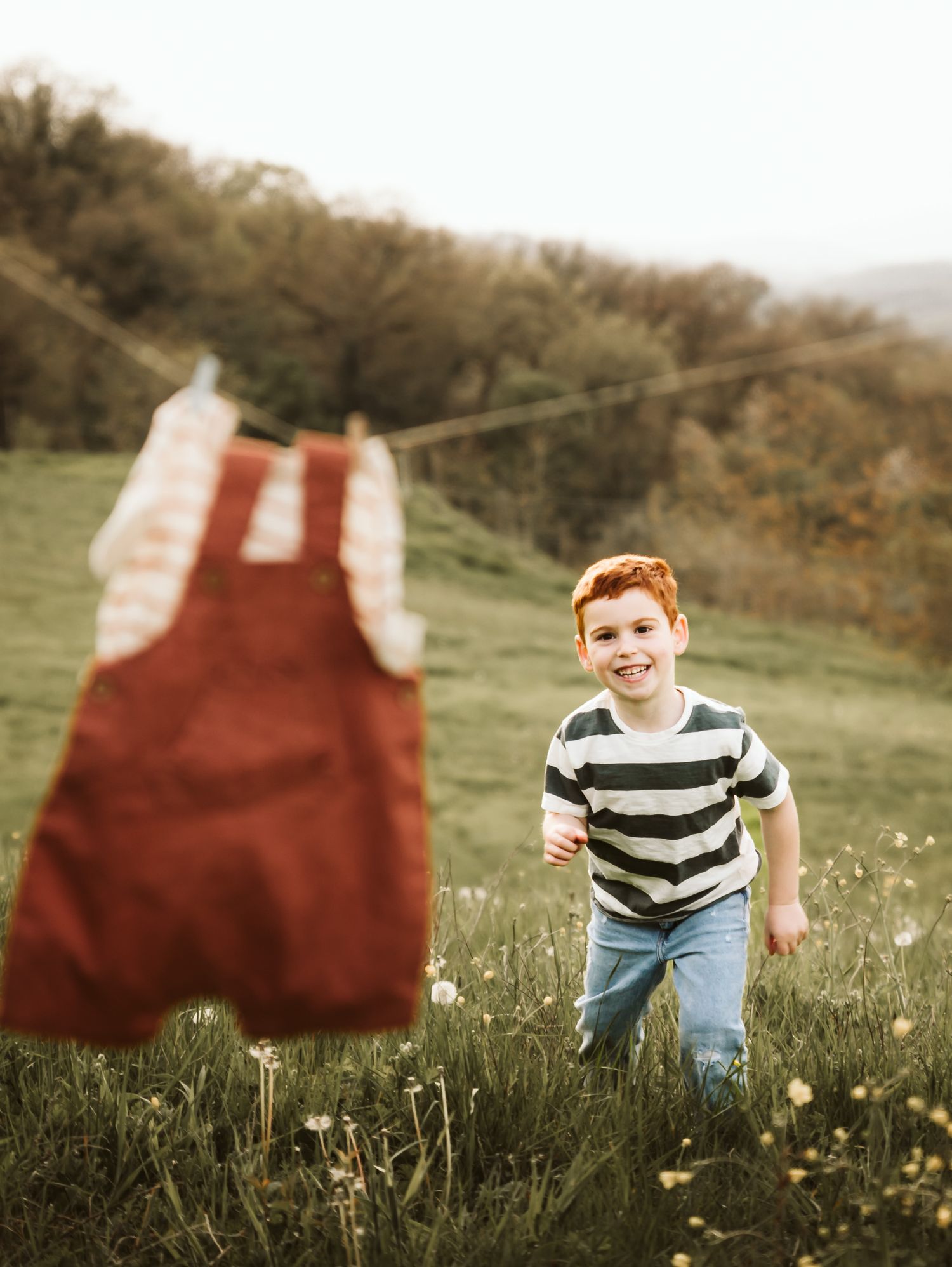 A happy red-haired boy in a striped shirt runs through a green meadow with clothes on a line.