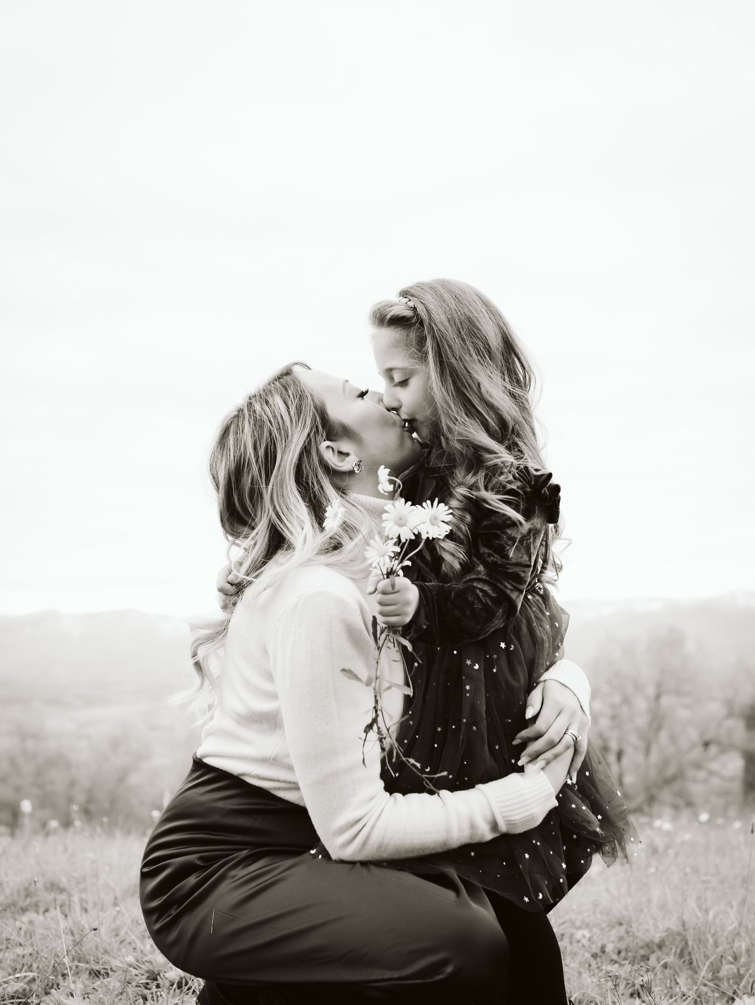 Black and white photo of mother kneeling to kiss her young daughter holding wildflowers outdoors.