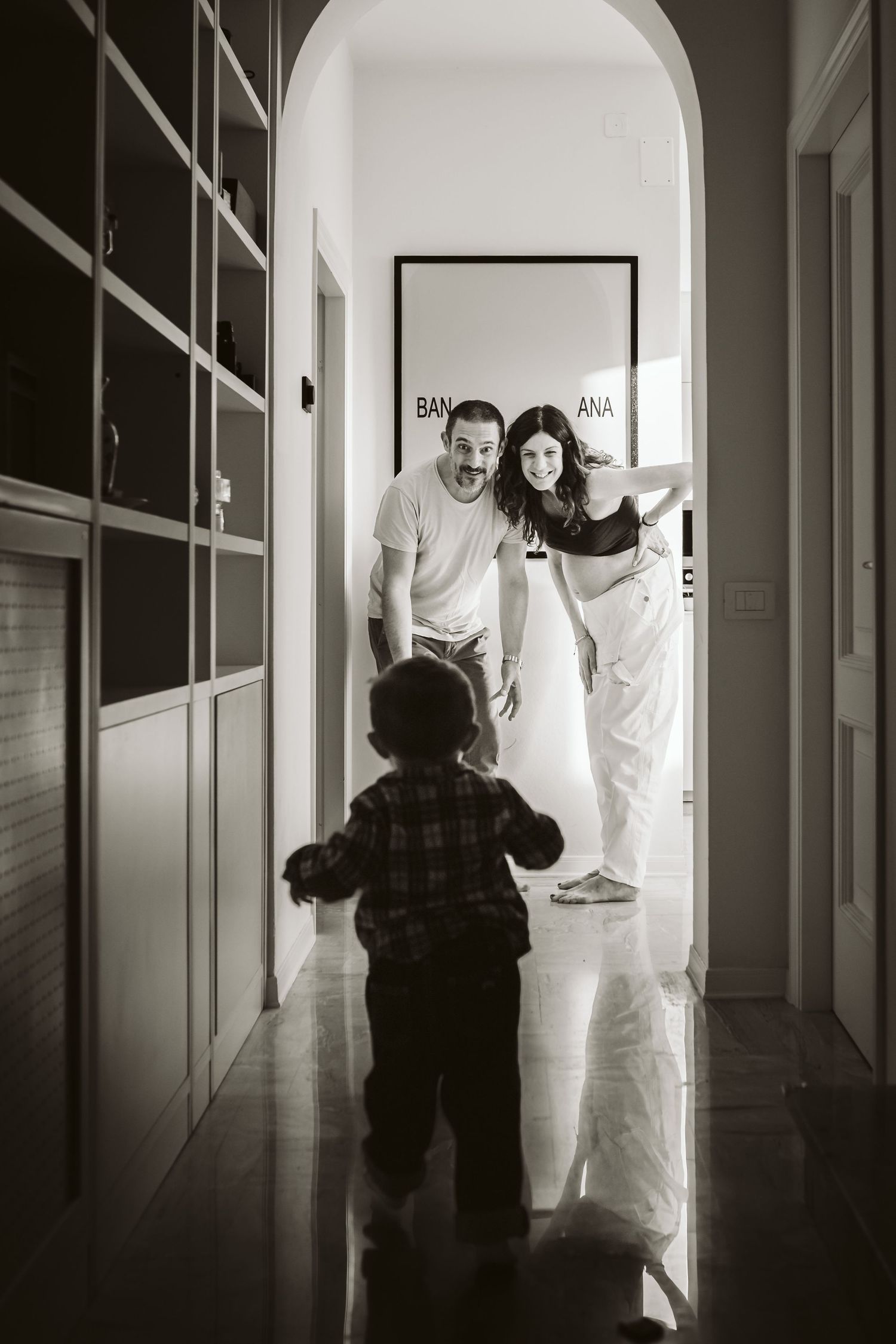 Black and white photo of toddler walking toward parents in a bright modern hallway.