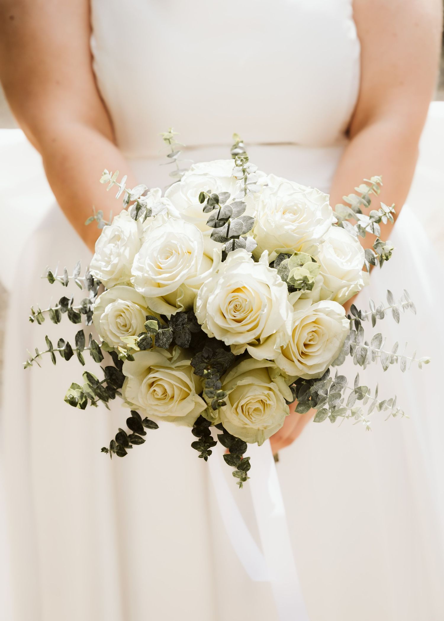 Bride holding elegant white rose and eucalyptus wedding bouquet in soft natural light.
