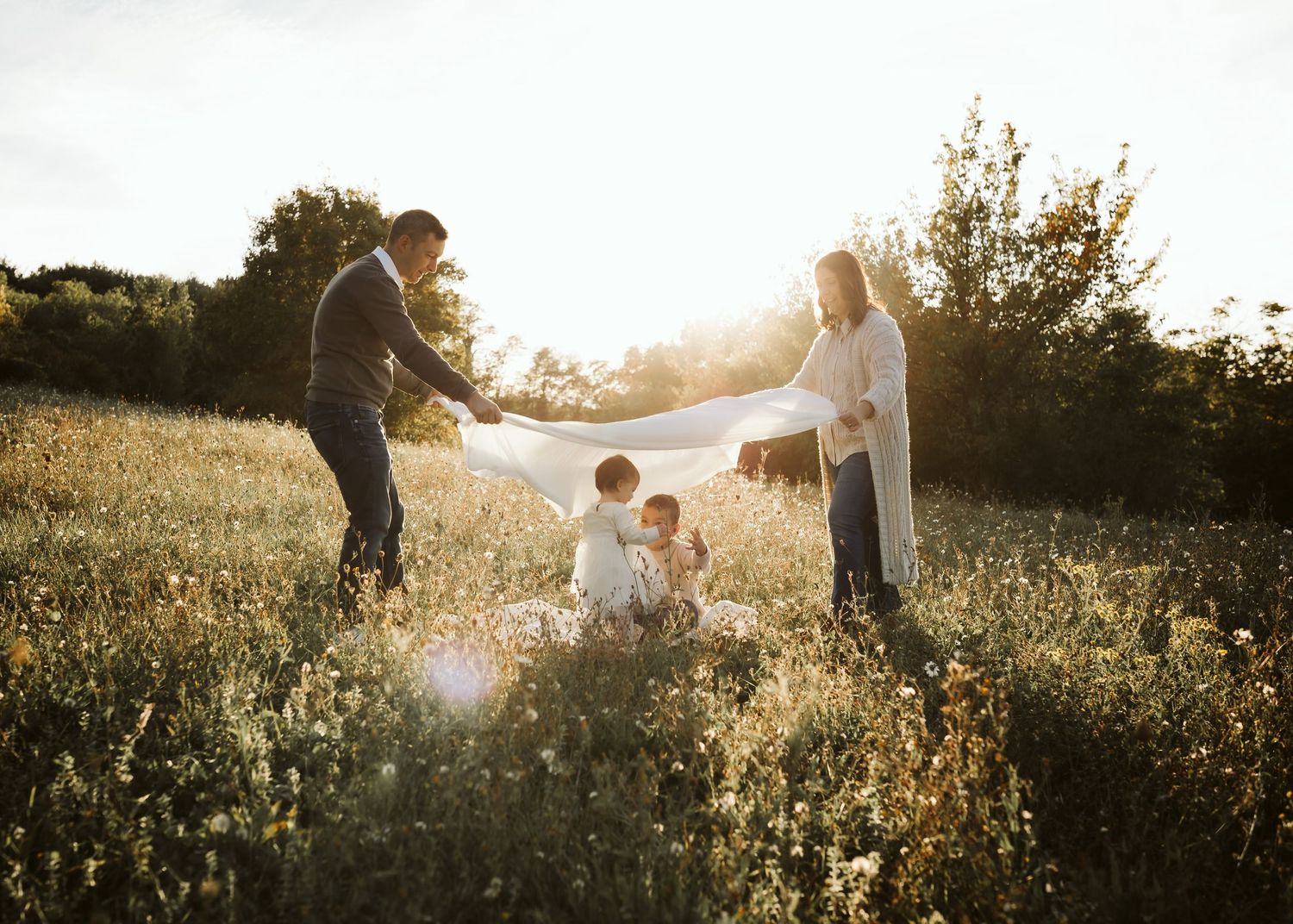 Family playing with white blanket in a sunny wildflower field.