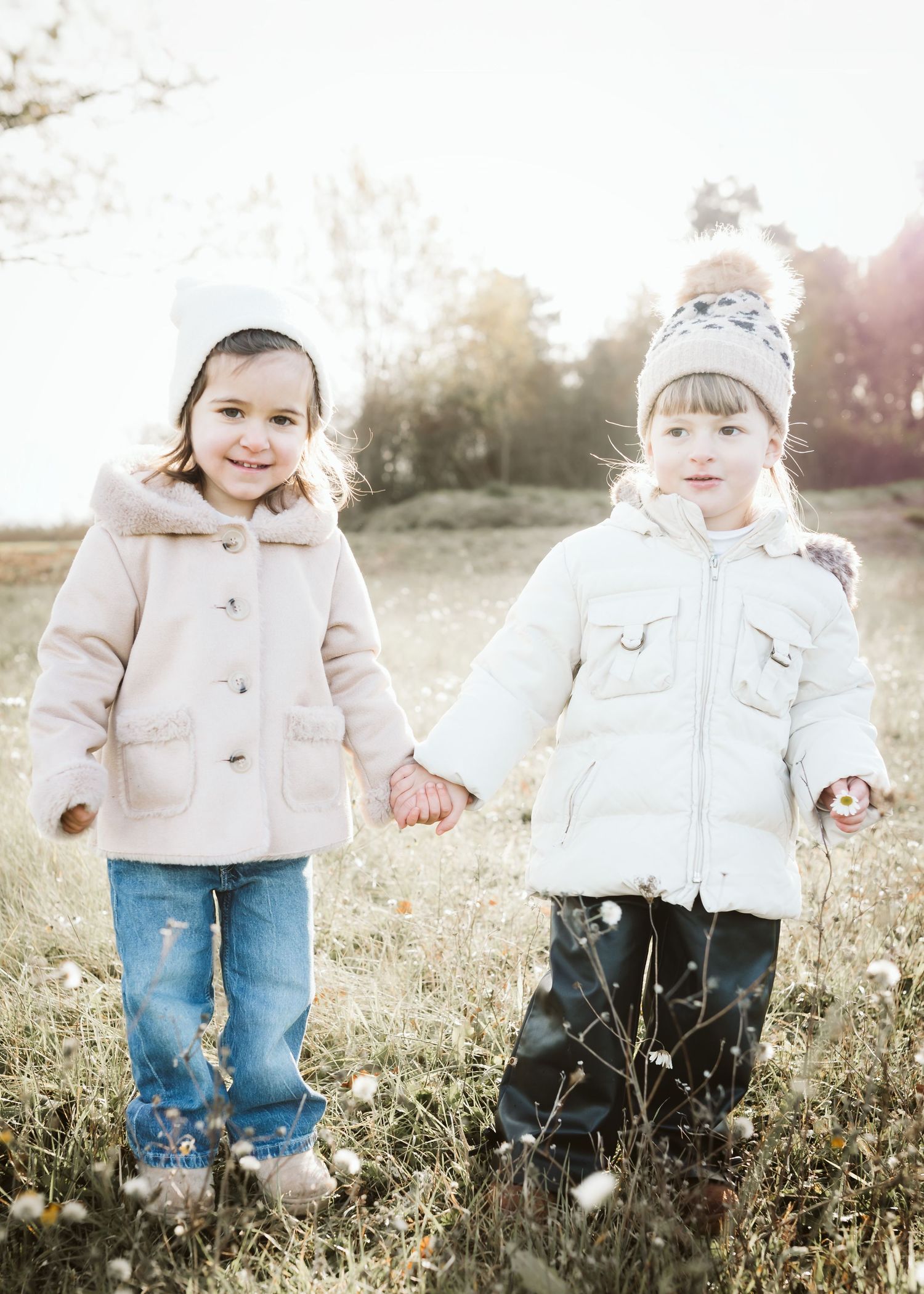 Two young girls in winter coats and hats holding hands in a sunny meadow.