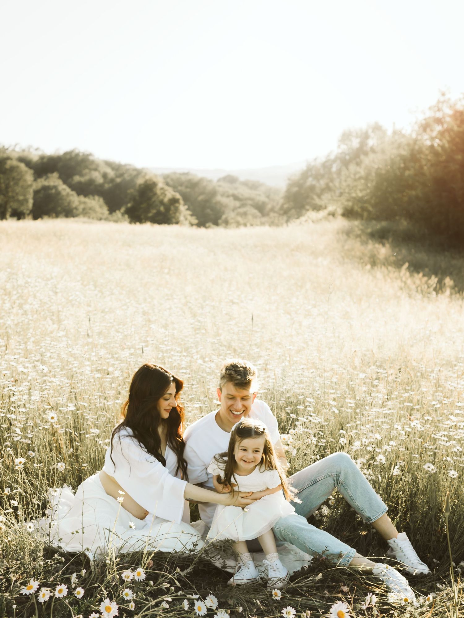 Happy family sitting in sunny daisy field, parents with young daughter in white outfits.