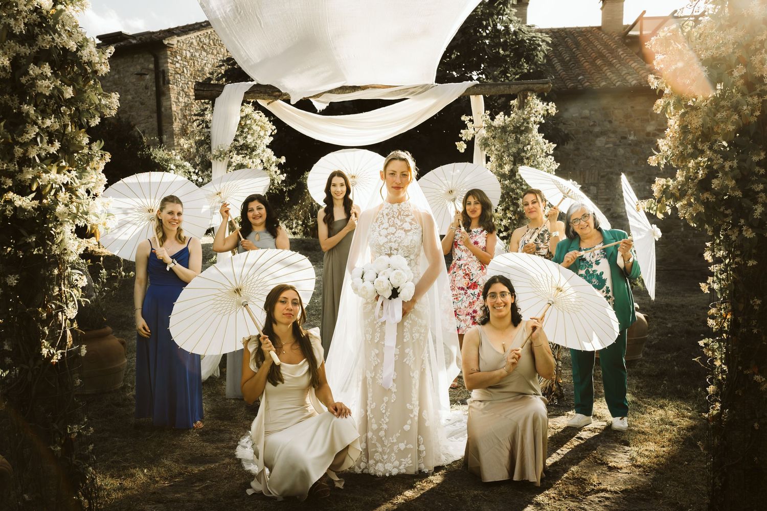 Bride surrounded by bridesmaids holding white parasols in sunlit rustic outdoor setting.