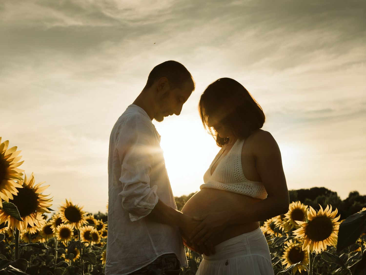 Couple silhouetted against golden sunset in a sunflower field, tenderly cradling a pregnant belly.