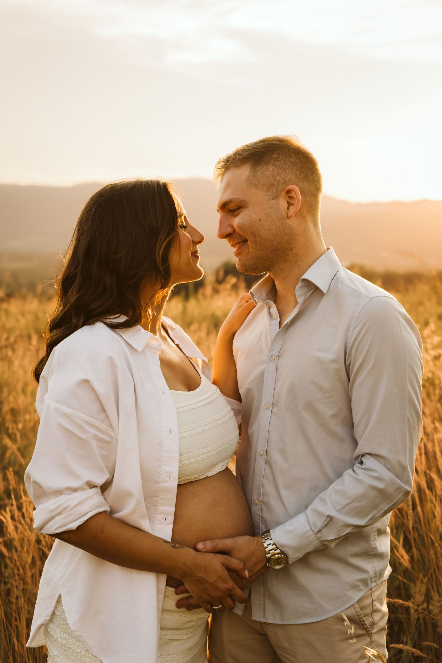 Expecting couple embracing in wheat field at sunset, hands on baby bump.