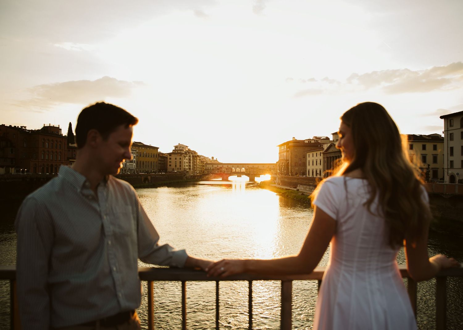 Couple holding hands at railing with Ponte Vecchio silhouetted against golden sunset.