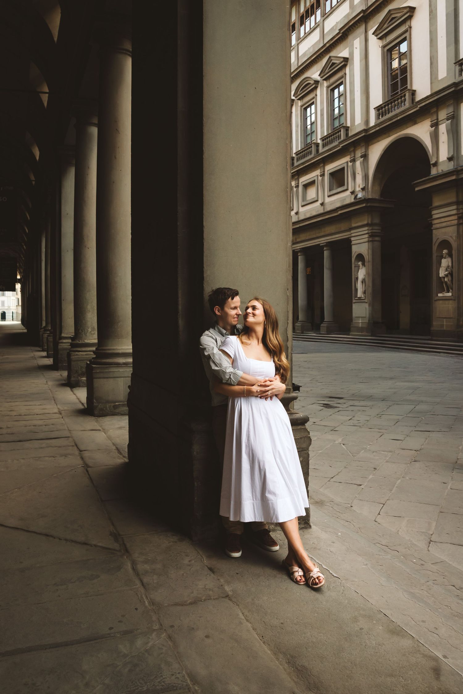 Couple embracing beneath stone columns in an elegant Italian Renaissance courtyard at golden hour.