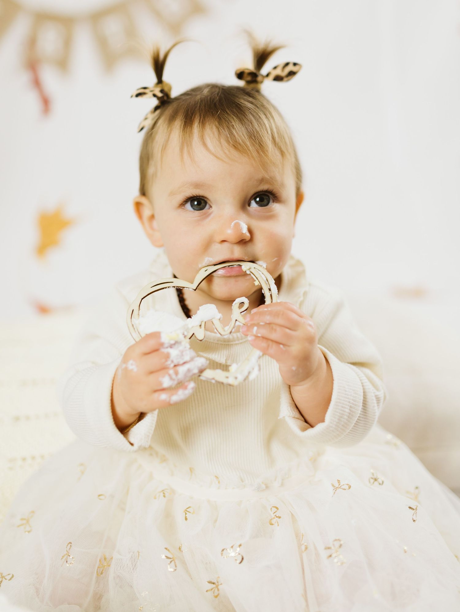 Adorable baby girl in white tutu dress chewing on a decorative number one at a birthday party.