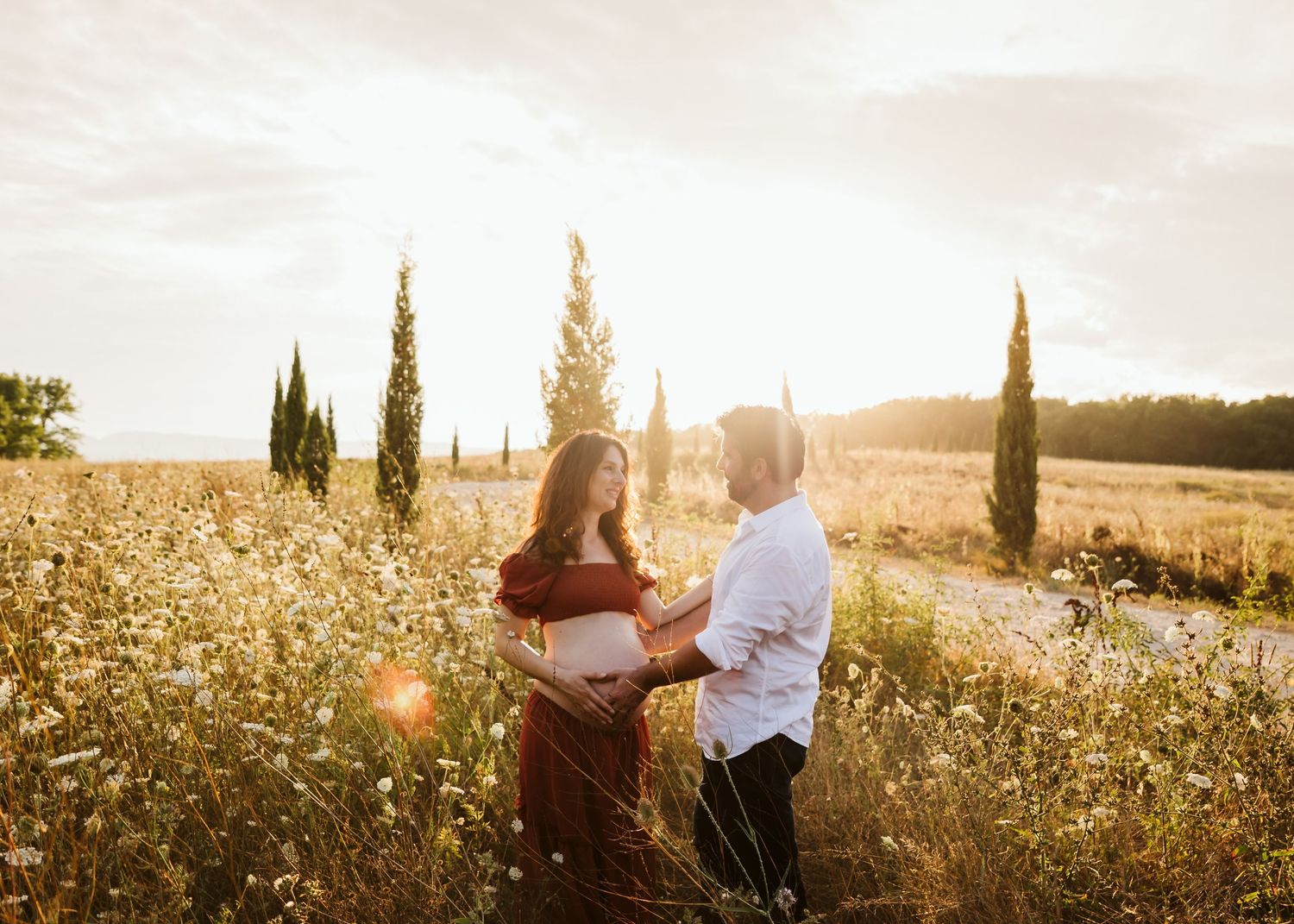 Pregnant couple holding hands in a sunlit wildflower field with cypress trees at golden hour.