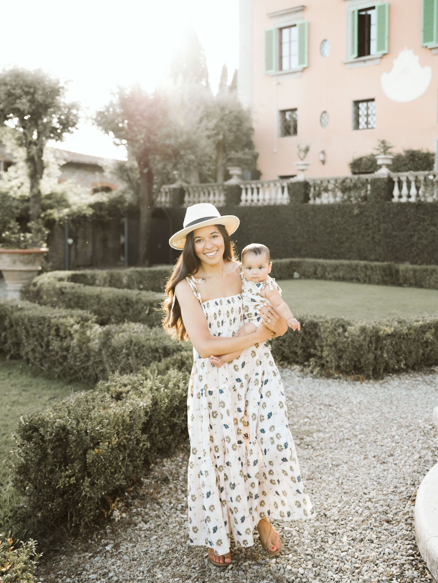 Smiling woman in floral dress and sun hat holding a baby in a manicured Italian villa garden.