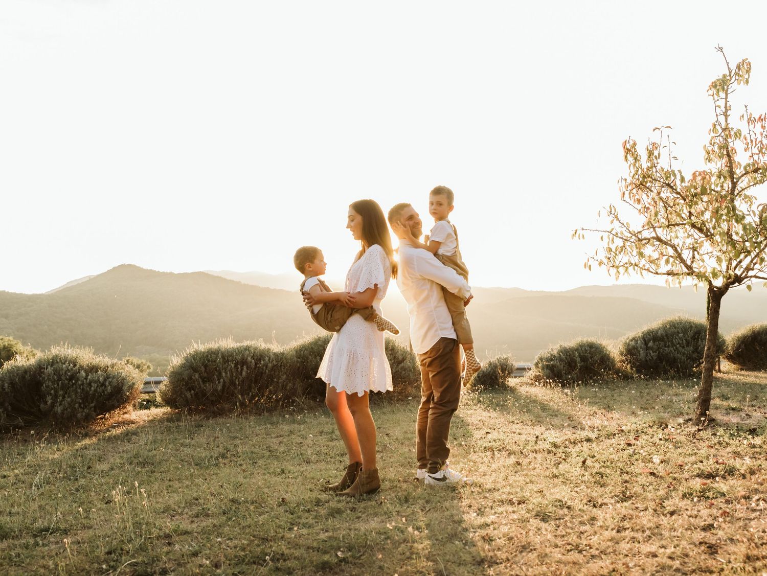 Family with two toddlers posing in a golden field with mountain views at sunset.