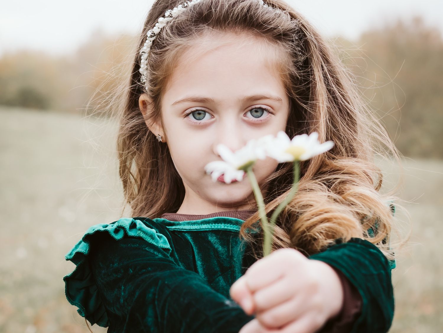 A young girl in a green velvet dress holds a white daisy flower toward the camera in an outdoor field.