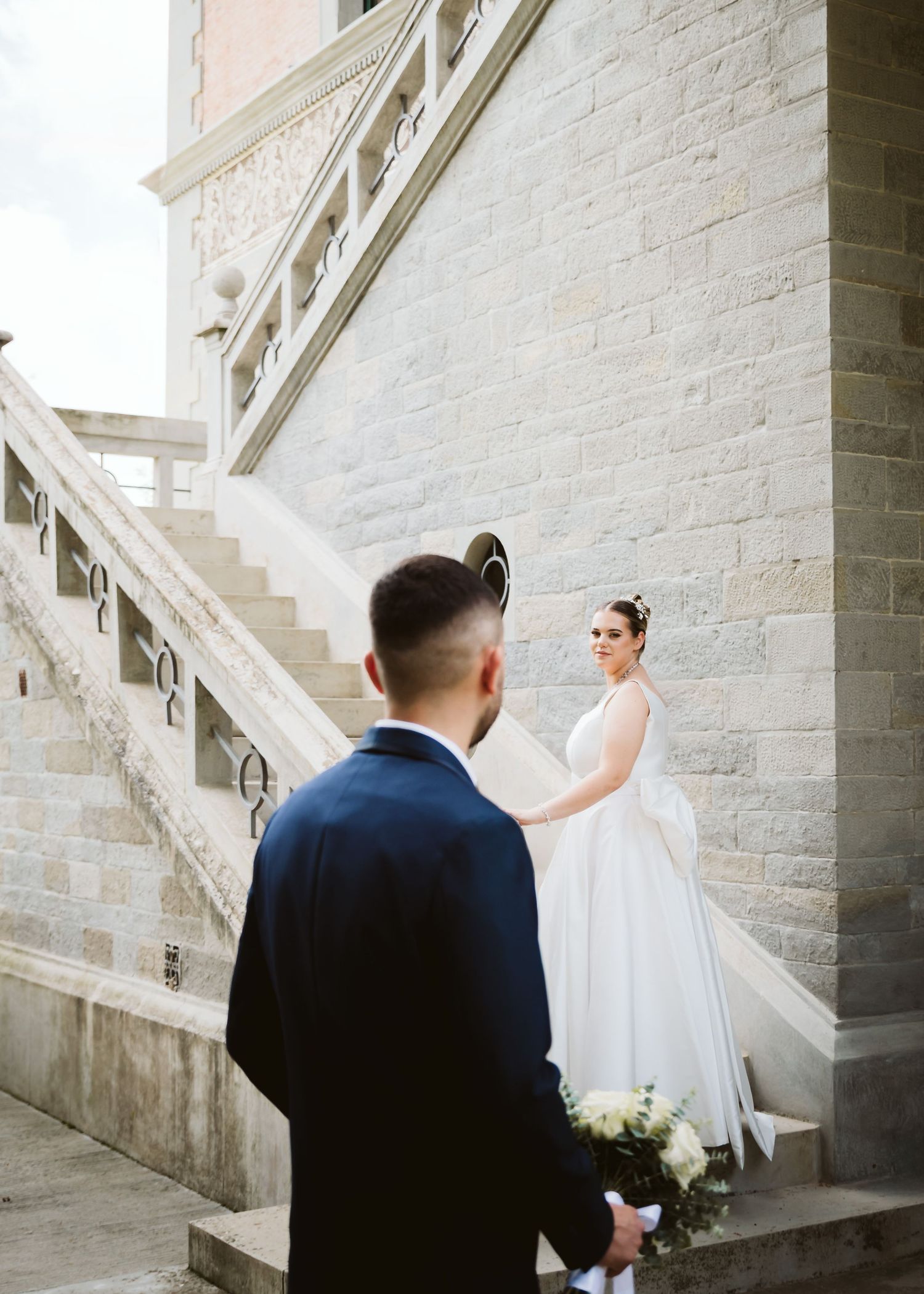 Bride in a white gown standing on stone stairs looking at groom holding a bouquet below.