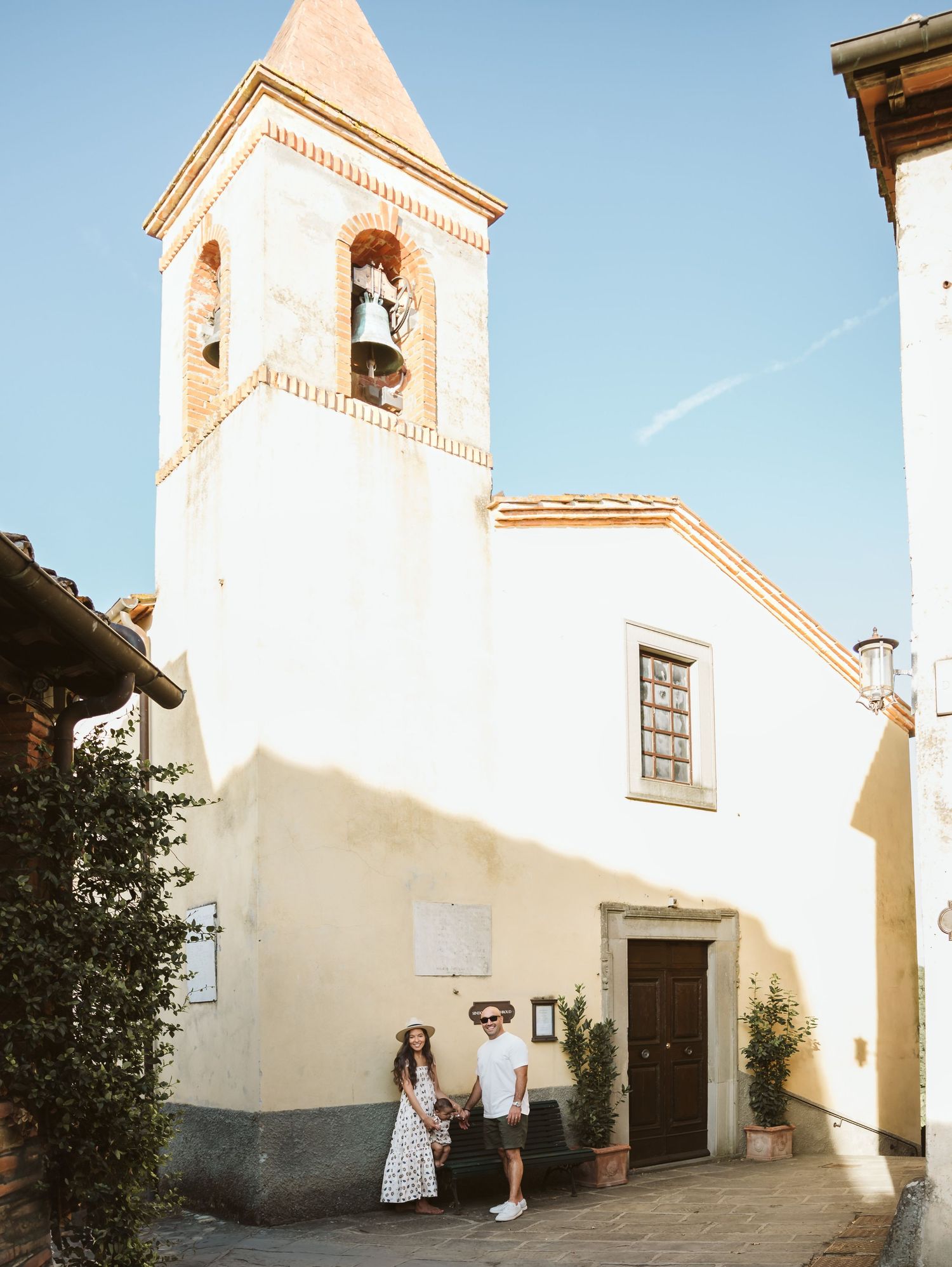Couple holding hands before a white Italian church with bell tower on sunny day.