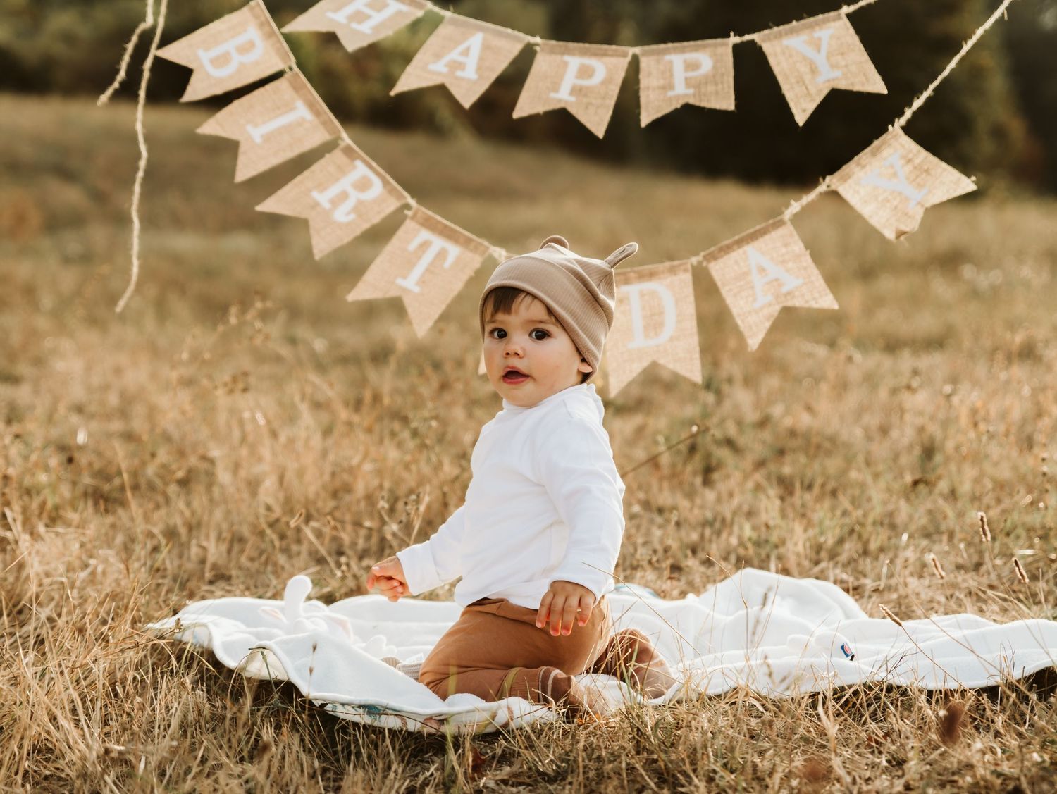 Adorable baby wearing a hat sits on a white blanket beneath a rustic Happy Birthday bunting banner.